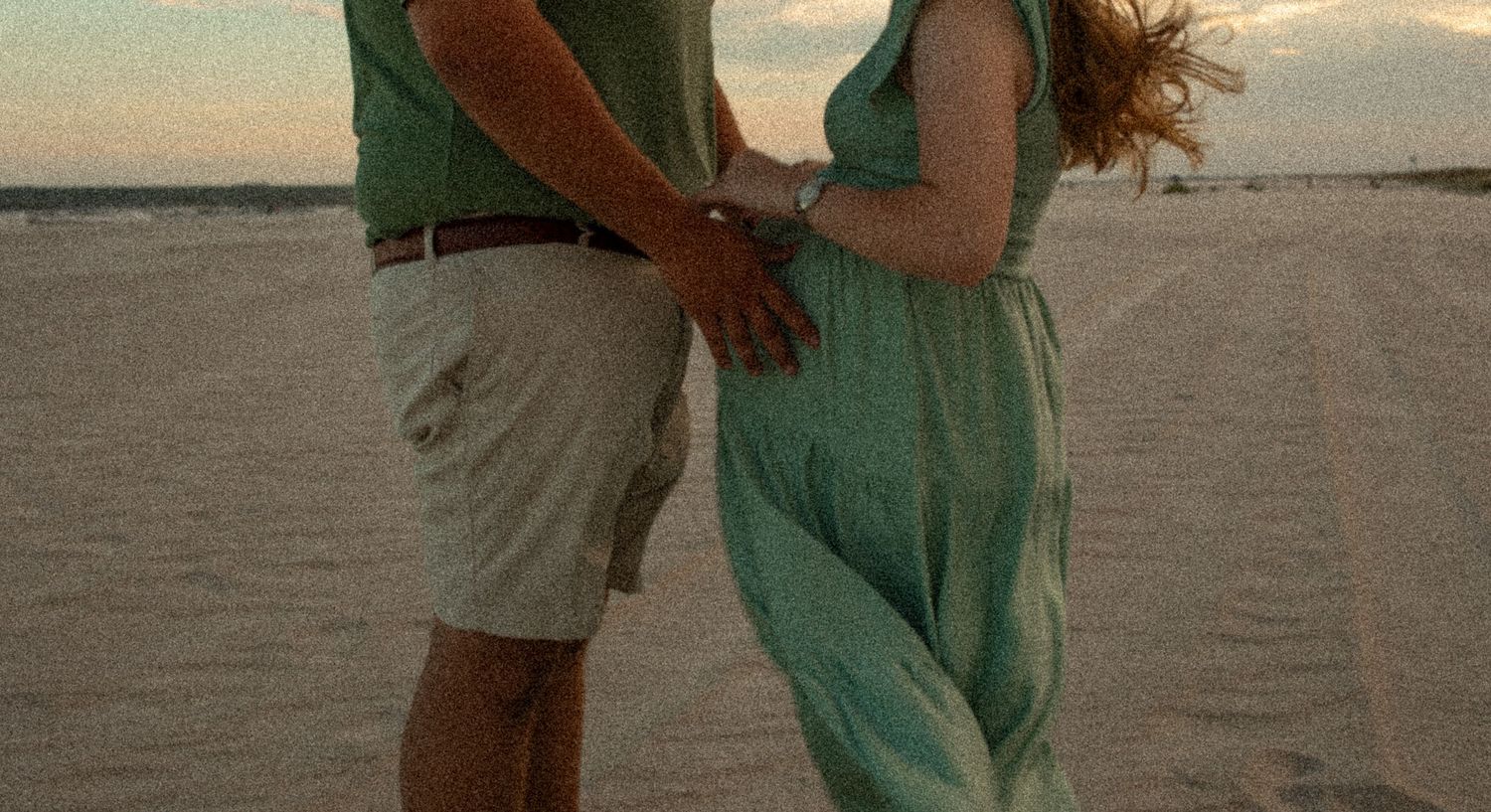 A couple holds hands on the beach at sunset in matching green and khaki attire.