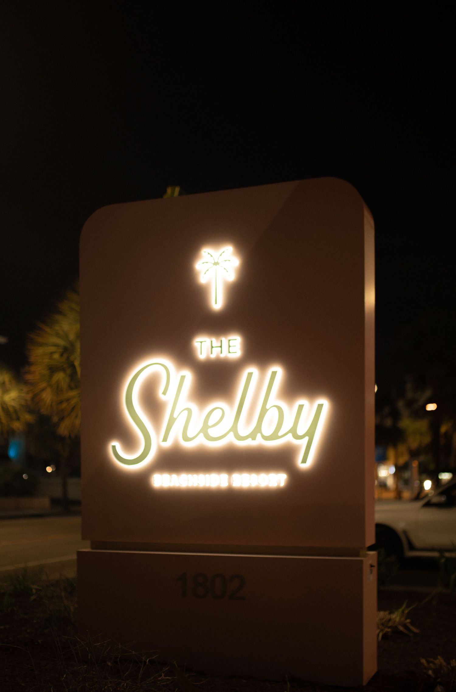 Illuminated sign for The Shelby Charming Resort at night, displaying address 1802, with glowing white and green lettering.