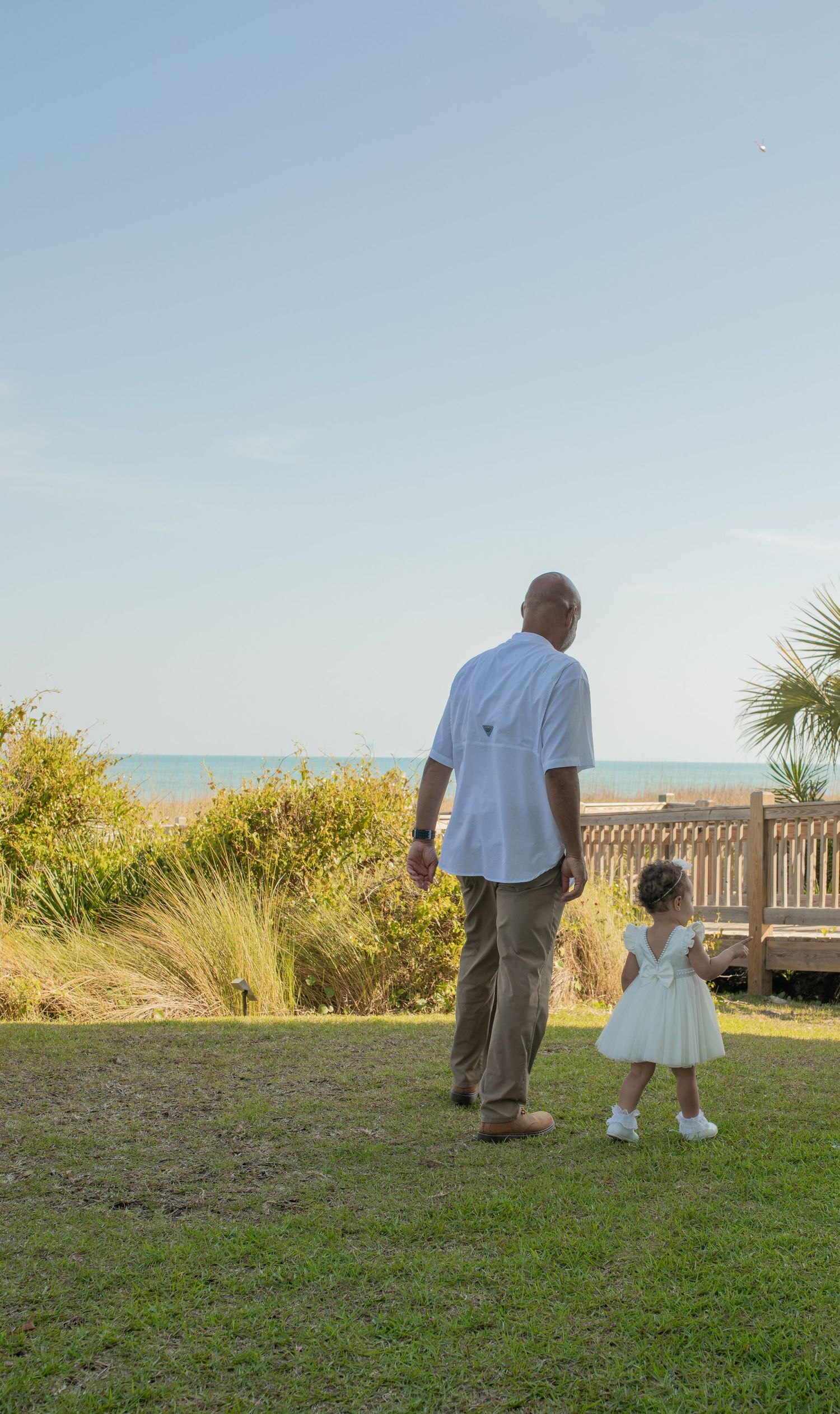 Man in white shirt holds hands with little girl in white dress walking toward beach boardwalk on sunny day.