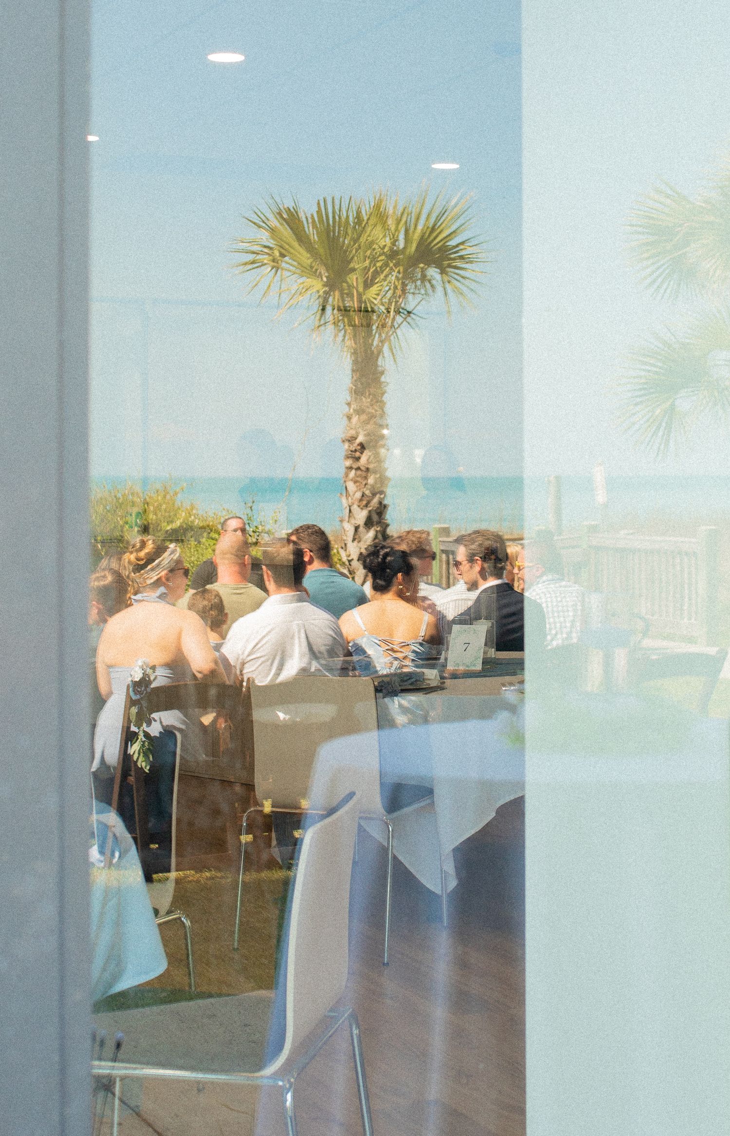 Guests gathered at an outdoor beachside event with palm trees and ocean views through a glass window.