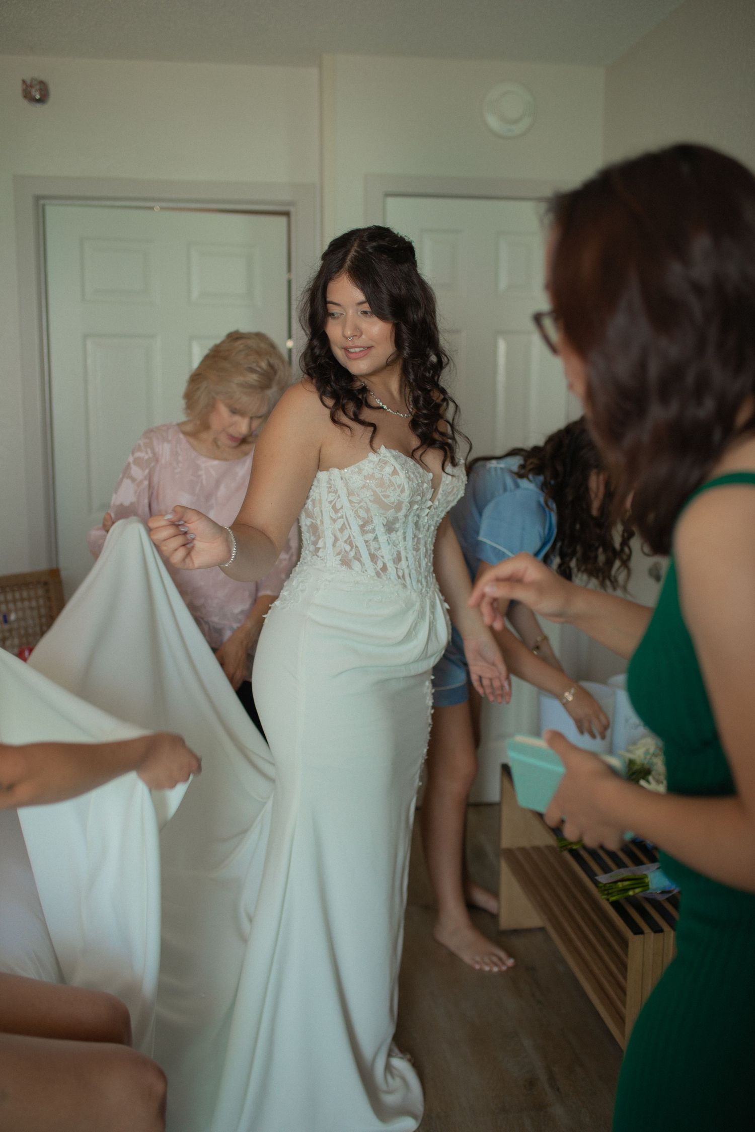 Bride in white lace wedding gown being assisted by bridesmaids while getting ready indoors.