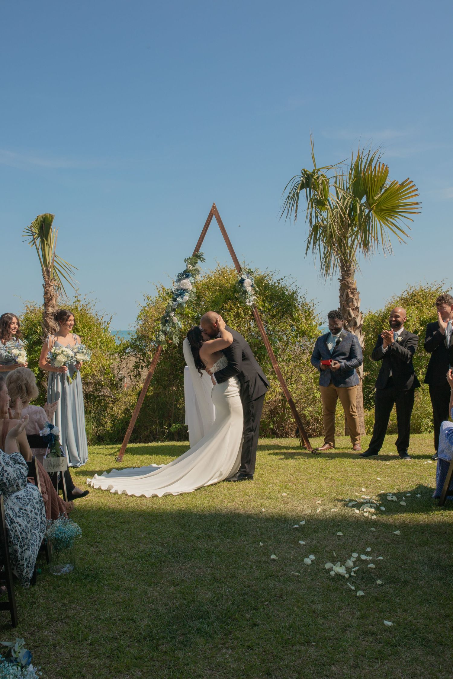 Bride and groom share first kiss under triangular arch at outdoor wedding ceremony with palm trees and guests.
