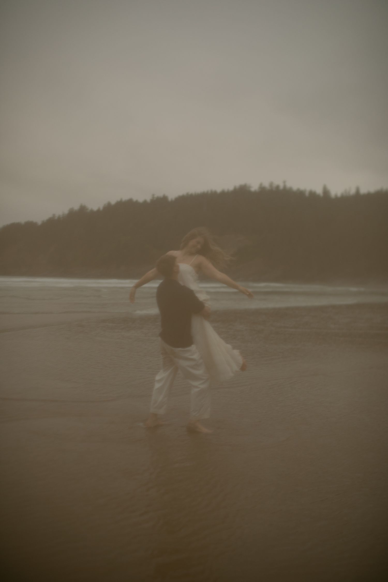 A couple dances barefoot on a misty beach in white attire, with forested hills and moody gray skies behind them.