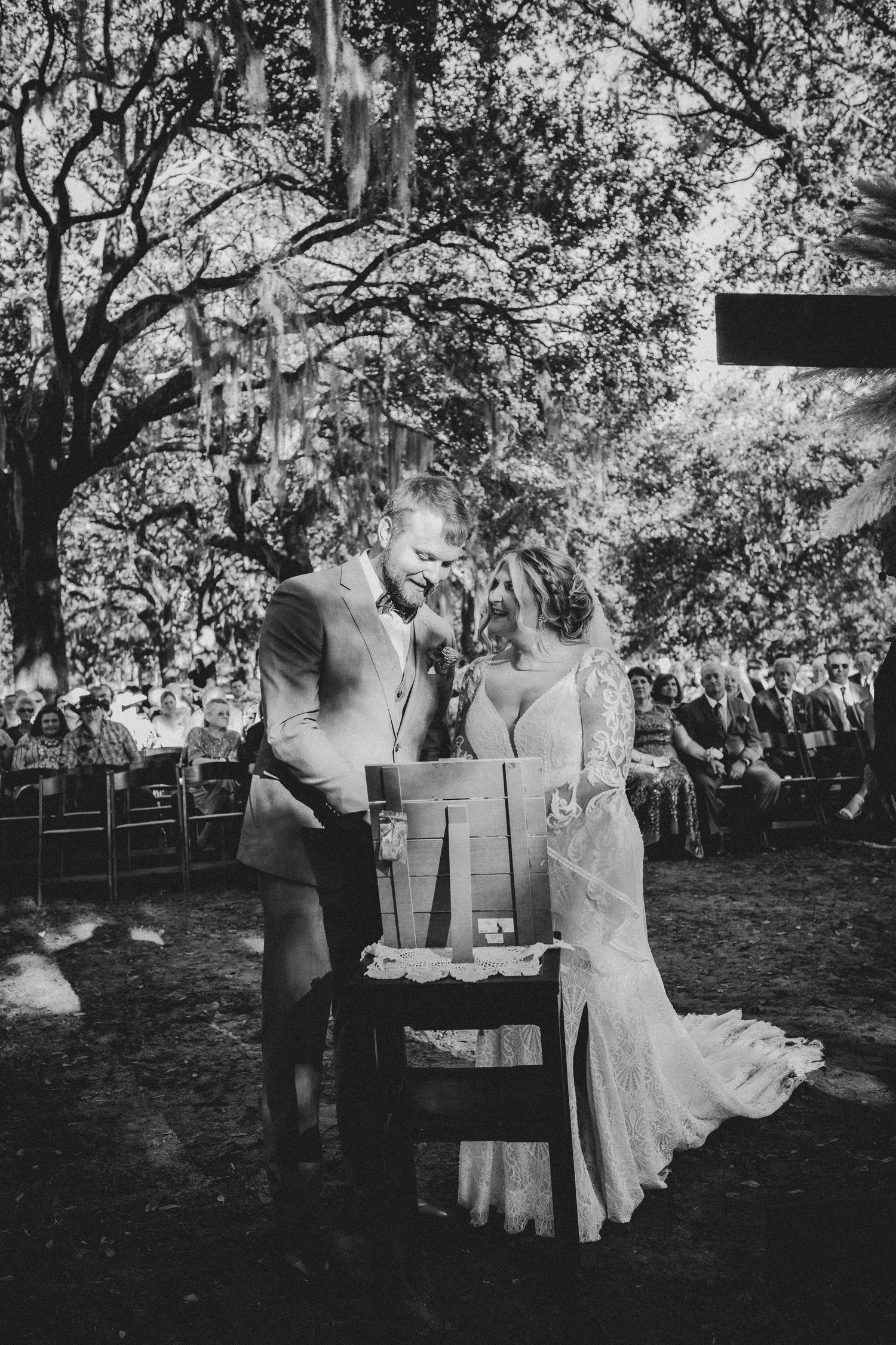 Black and white photo of a wedding ceremony outdoors with couple standing at a rustic wooden table under trees.