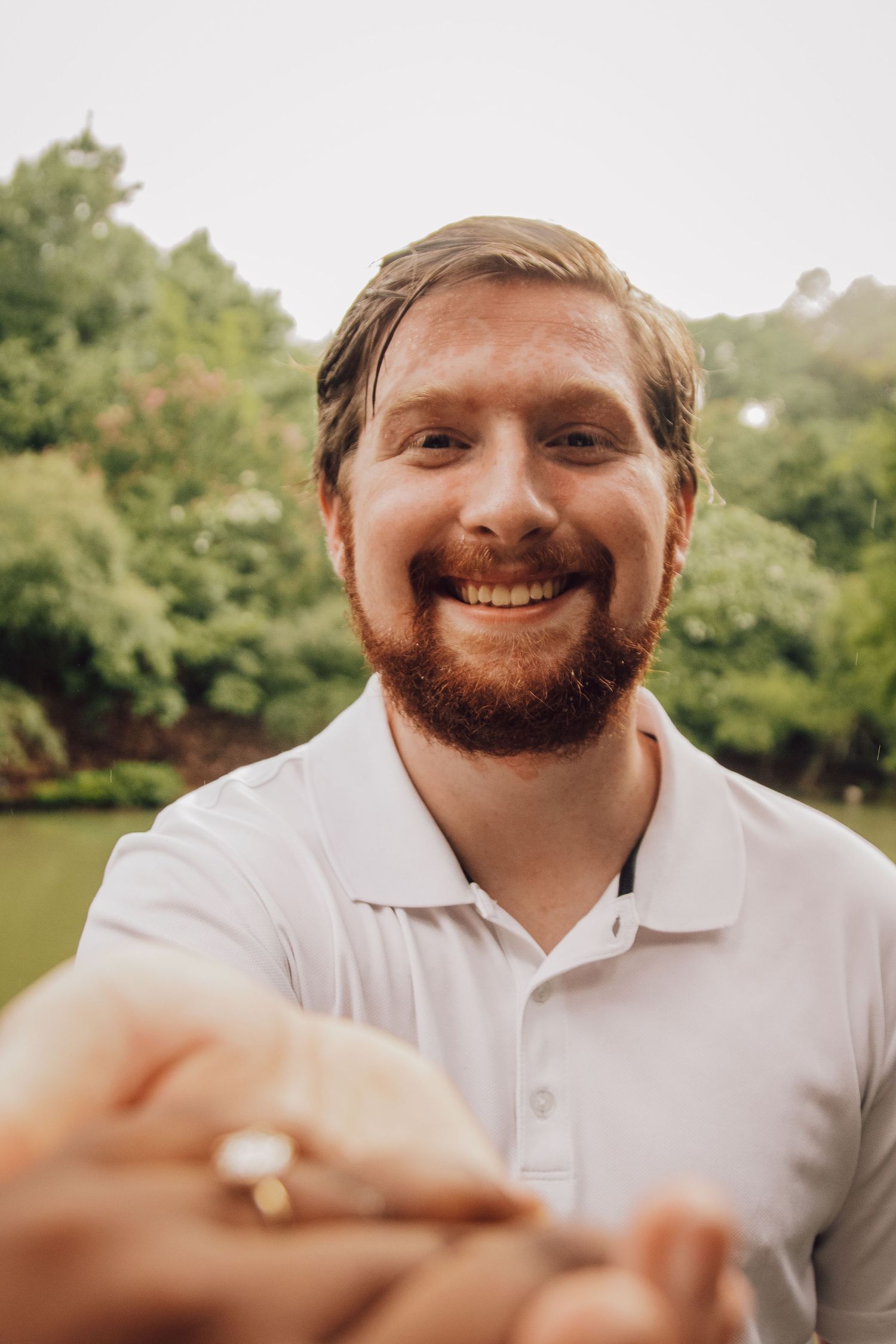 Smiling bearded person in white polo shirt posing outdoors with greenery in background.