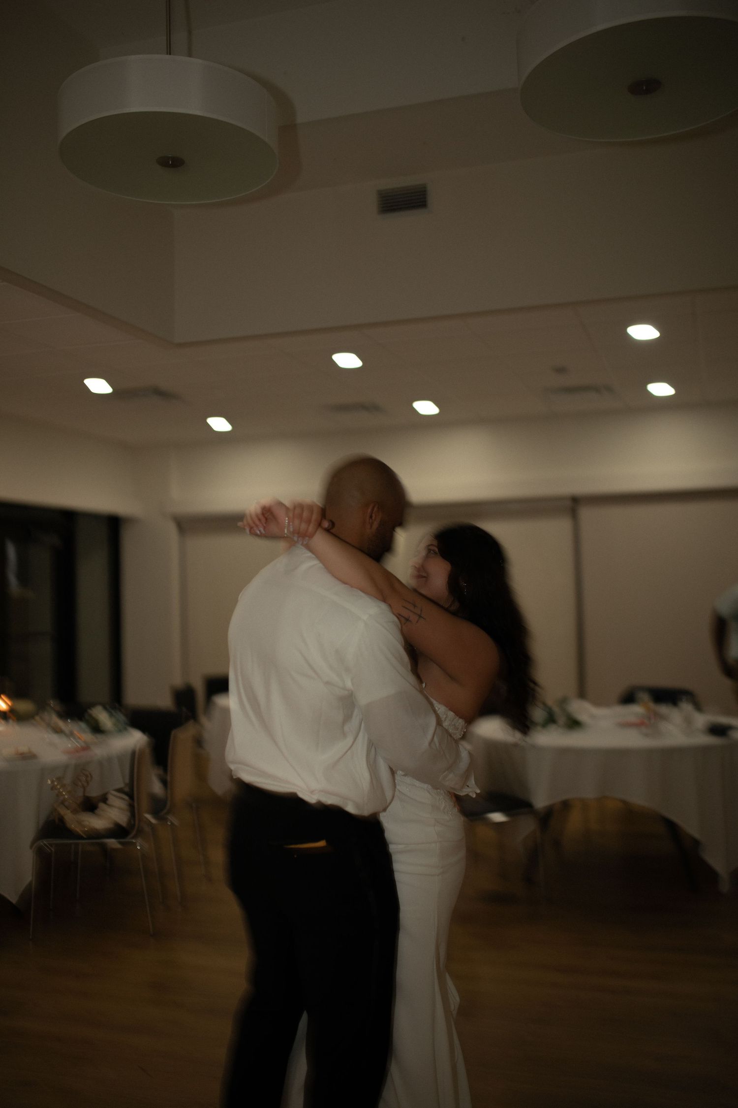 A couple shares their first dance in a softly lit wedding reception hall, the bride in white and groom in a dress shirt.