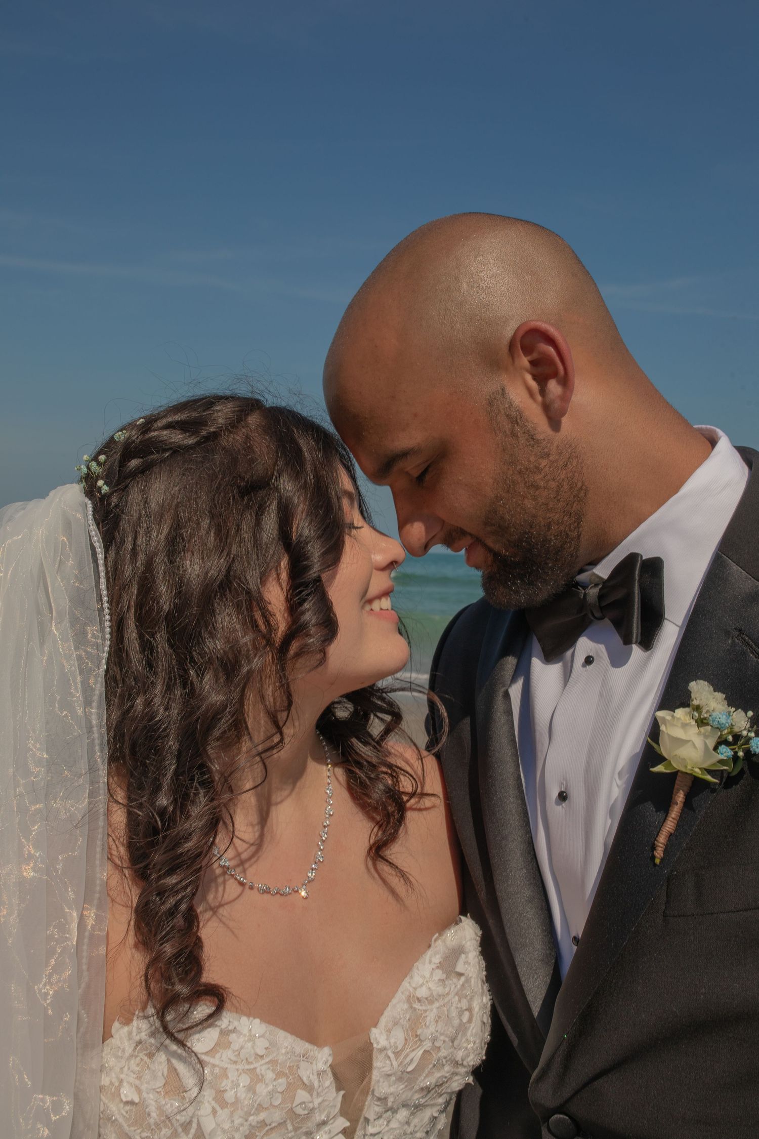 Bride and groom share a romantic forehead touch on a beach, wearing wedding attire with ocean backdrop.