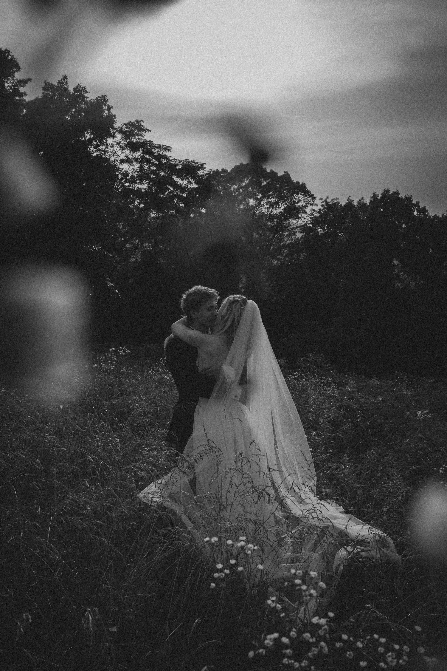 Black and white photo of a bride and groom kissing in a wildflower meadow with trees in the background.