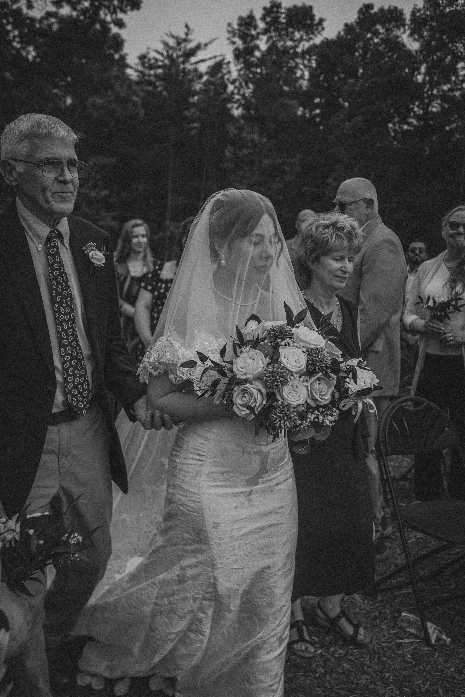 Black and white photo of a bride in veil holding bouquet, escorted down the aisle by her parents at an outdoor wedding.