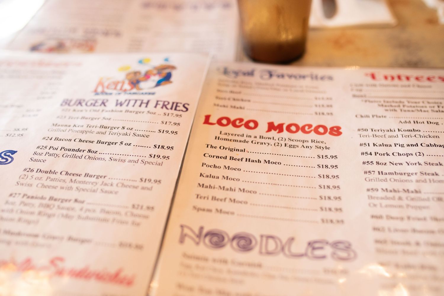 Close-up view of a restaurant menu showing sections like Loco Mocos and Noodles with prices listed in soft focus.