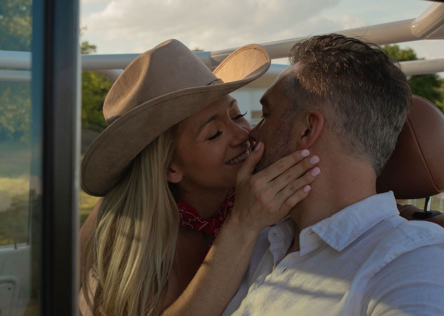 A blonde woman in a cowboy hat shares a romantic kiss with a gray-haired man in an outdoor setting.