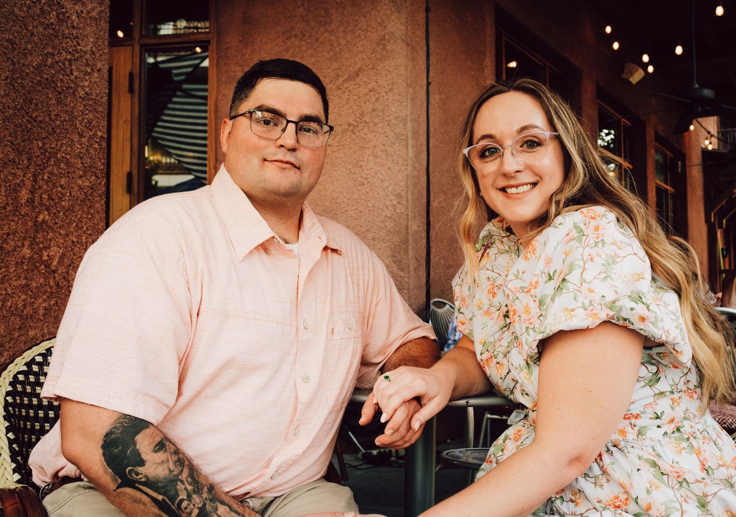 A couple seated at an outdoor cafe smiling in coordinated pink and floral outfits.