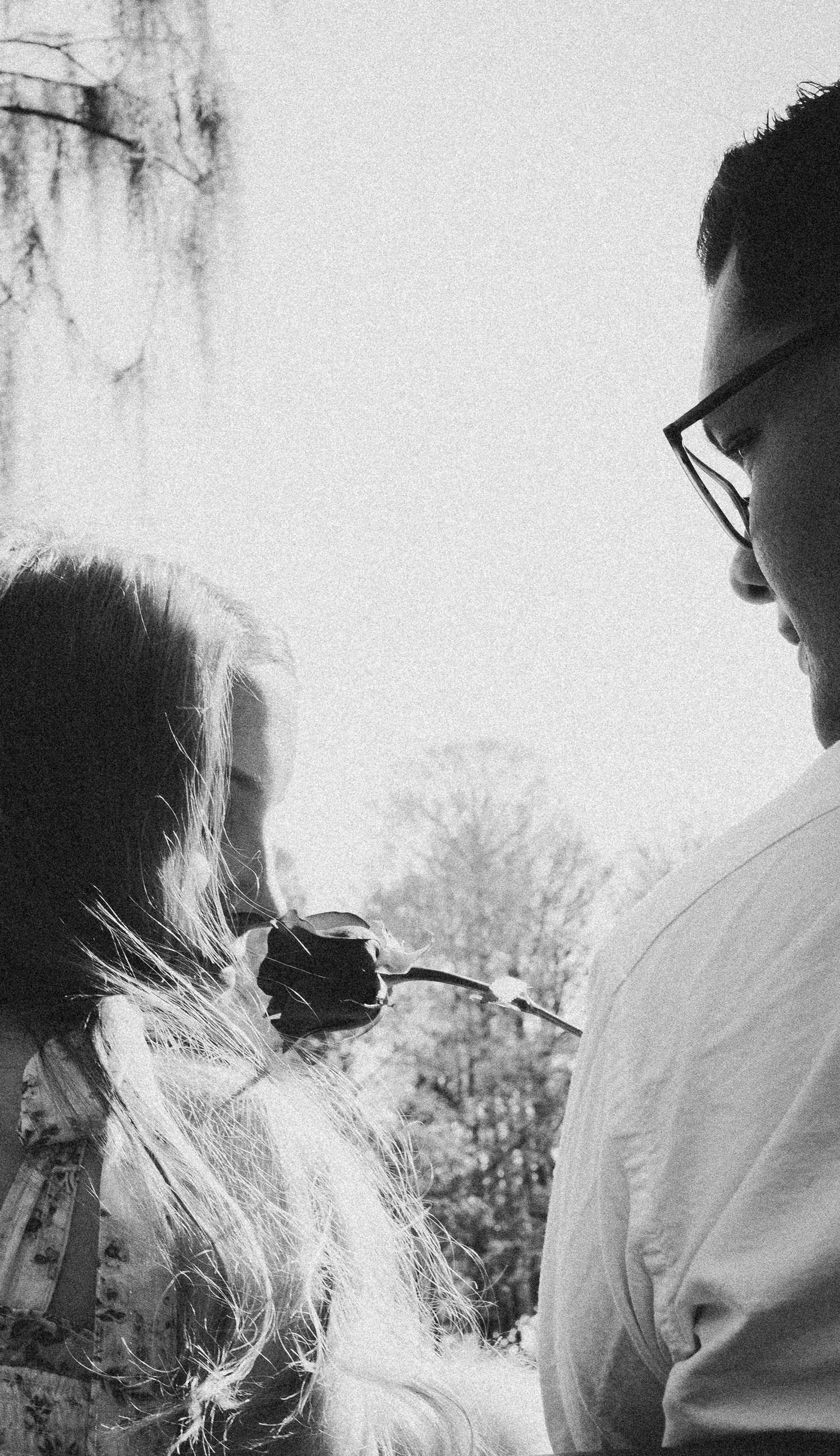 A black and white photo showing a side profile wearing glasses while holding a rose outdoors.