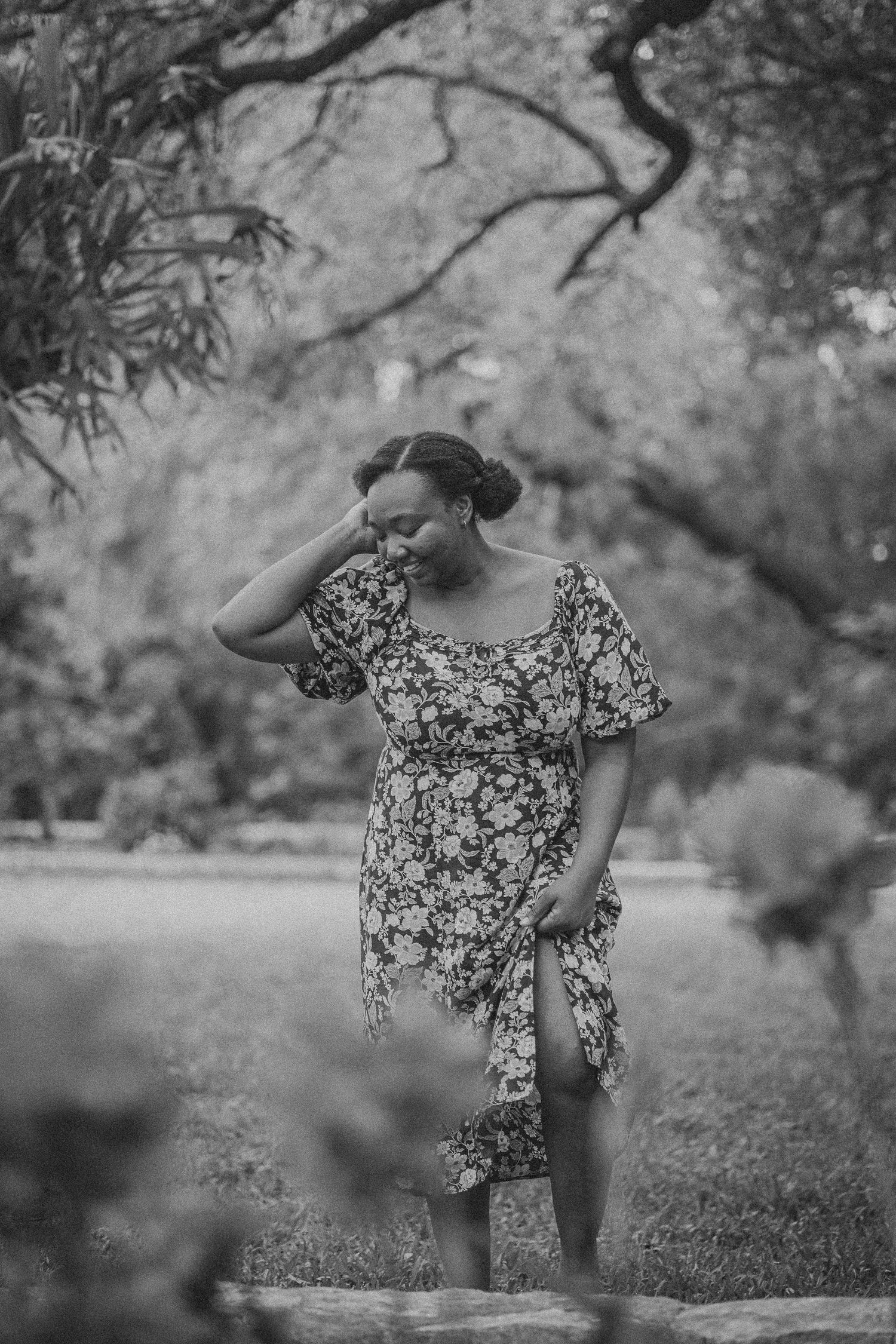 Black and white photo of a woman in a floral dress posing gracefully in a lush green park setting.