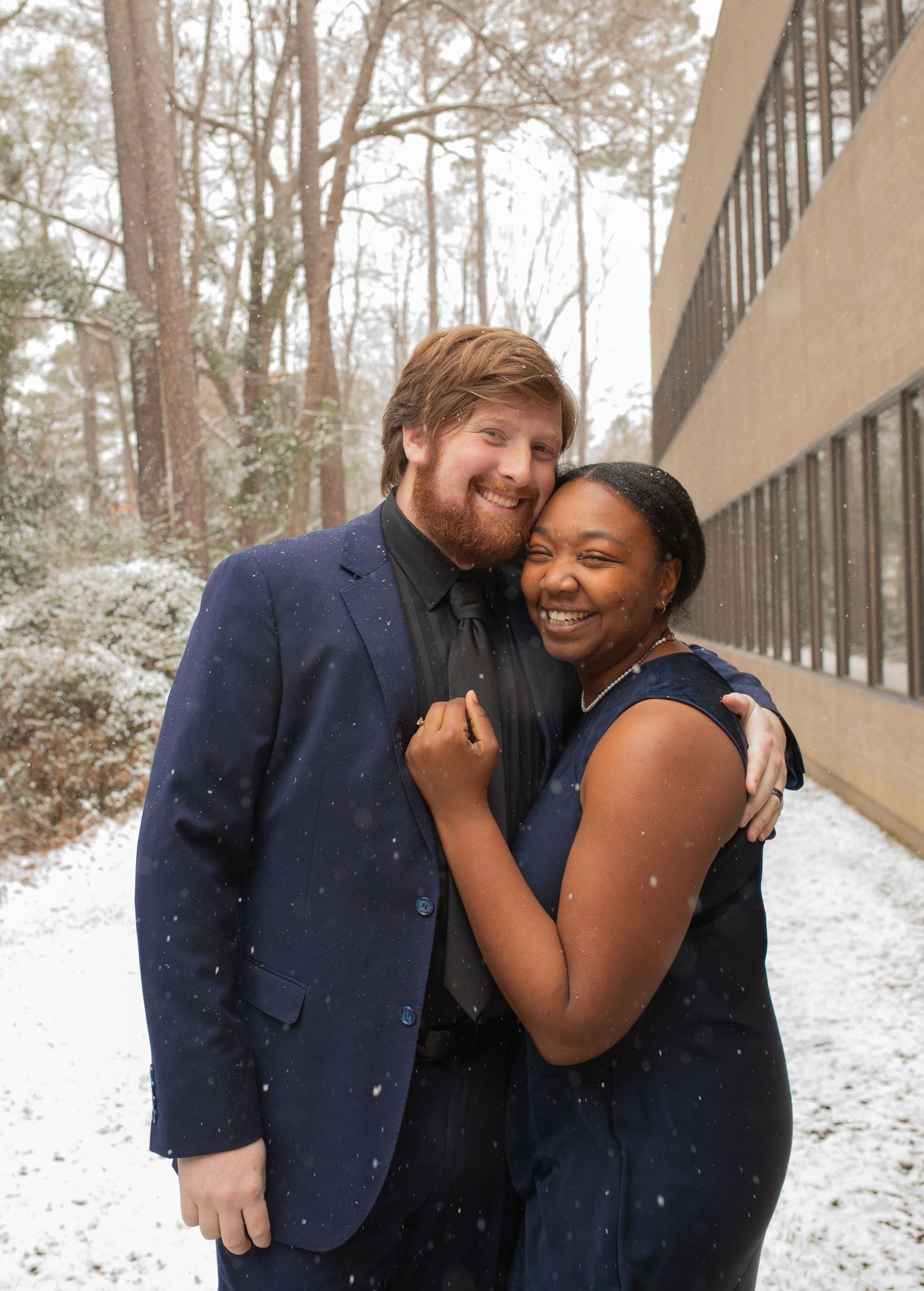 Couple embracing and smiling in light snowfall outdoors, wearing navy blue formal attire, with snowy trees in background.