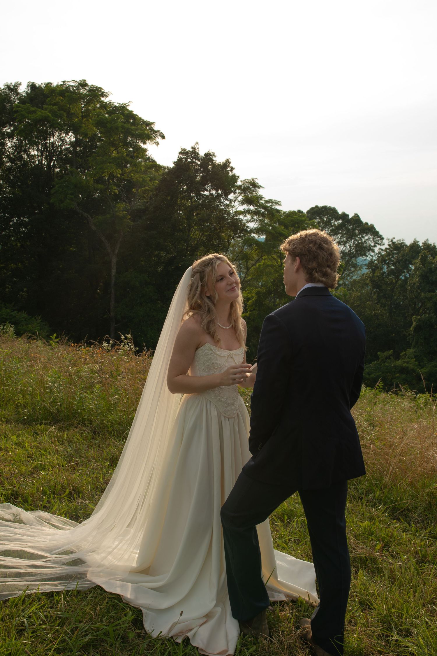Bride in white gown and veil stands with groom in navy suit in a lush green meadow during golden hour.