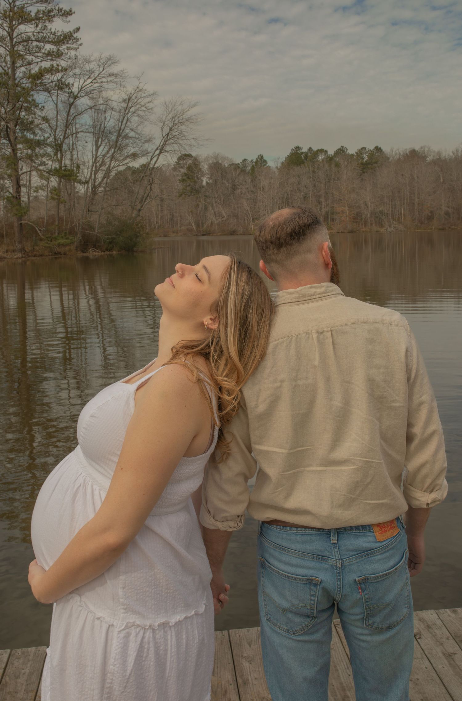 Pregnant woman in white dress leans on man in beige shirt on dock by serene lake surrounded by trees.