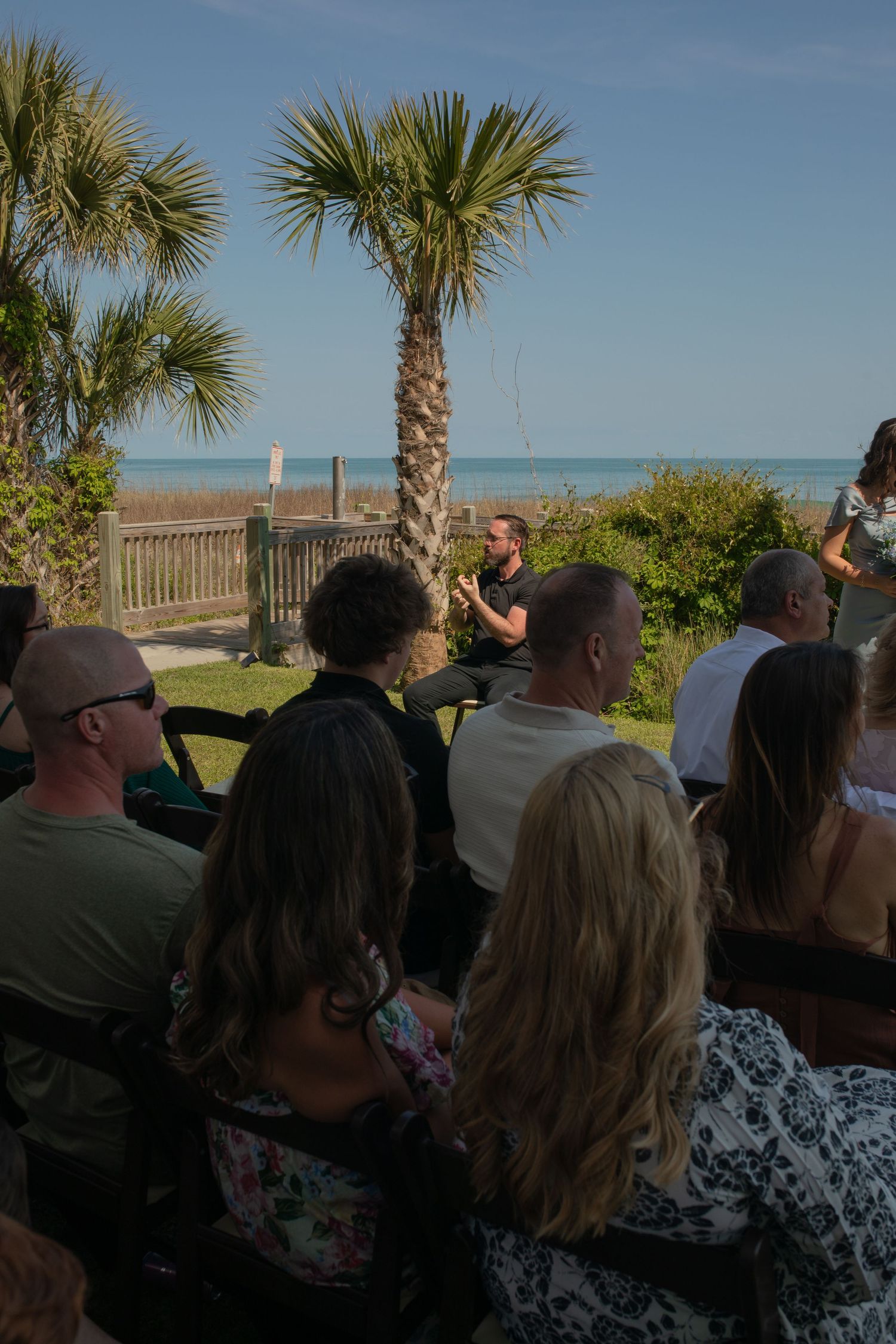 Outdoor beachside gathering with audience seated facing a speaker near palm trees and ocean views.