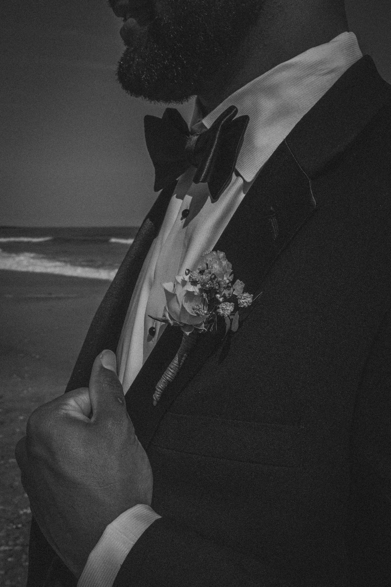 Black and white photo of a groom in a tuxedo with boutonniere and bow tie at a beach wedding.