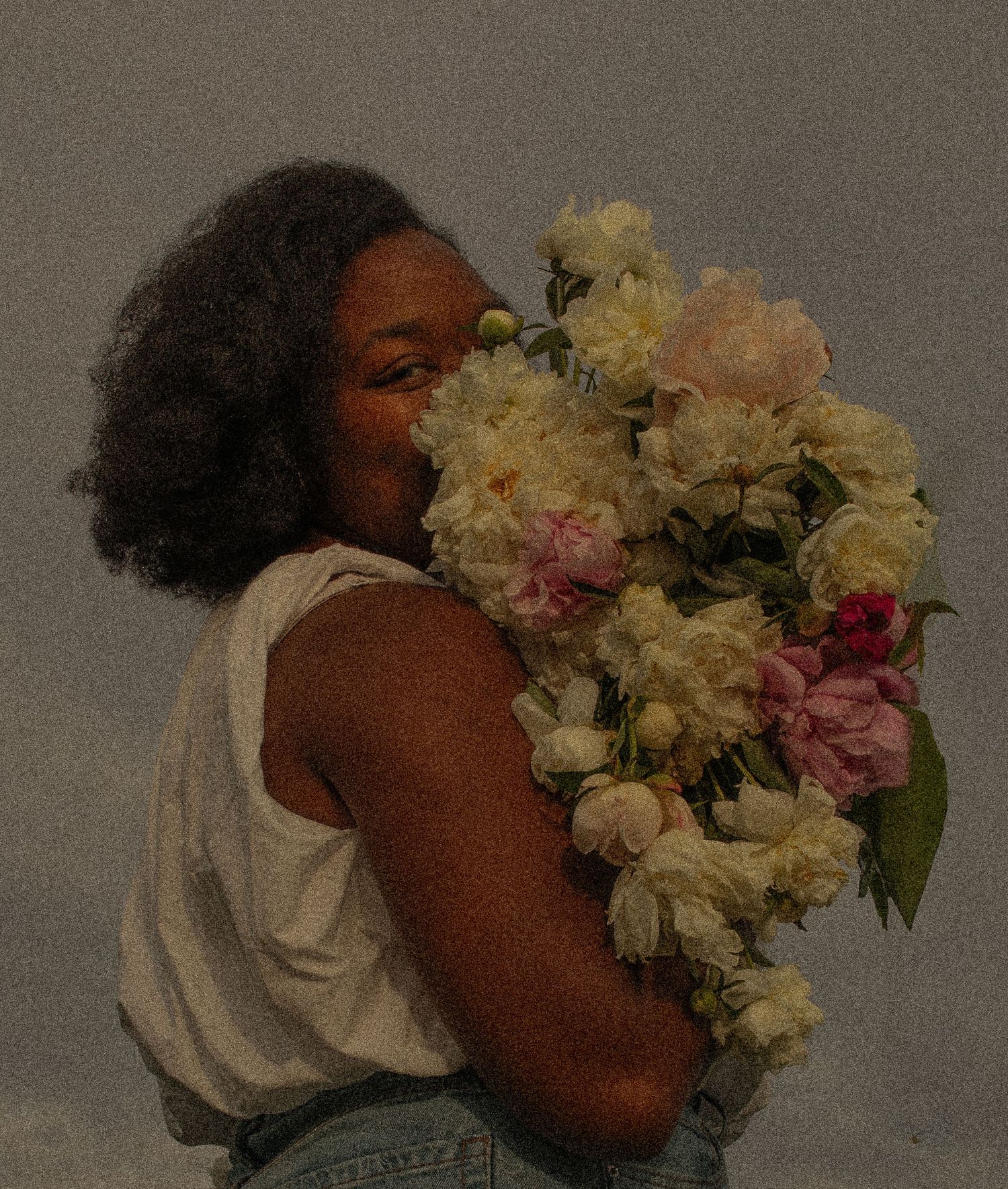 A person in a white tank top holds a lush bouquet of pink and cream peonies against a dark background.