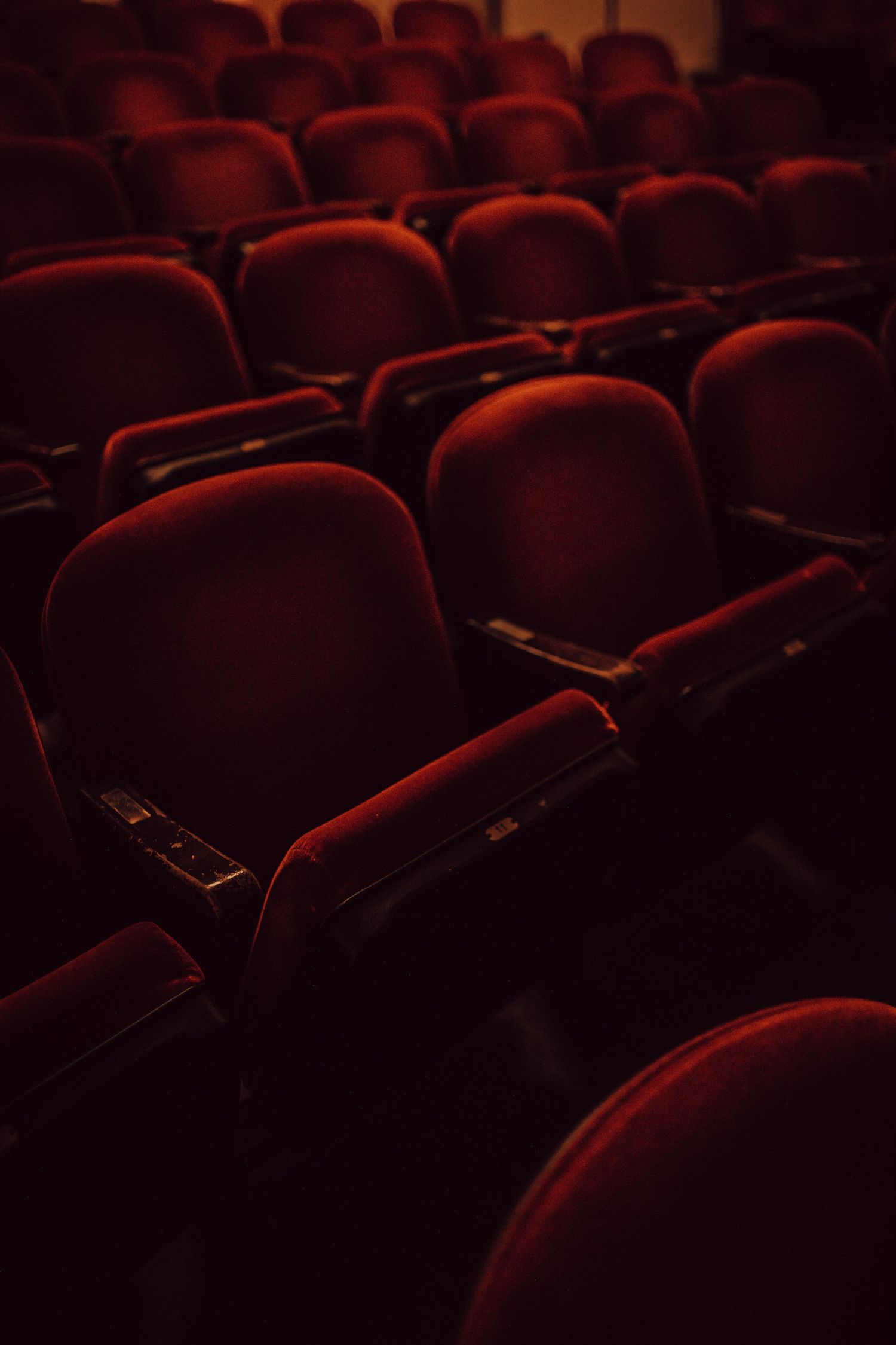 Empty theater seats in deep red velvet arranged in symmetrical rows under dramatic lighting.