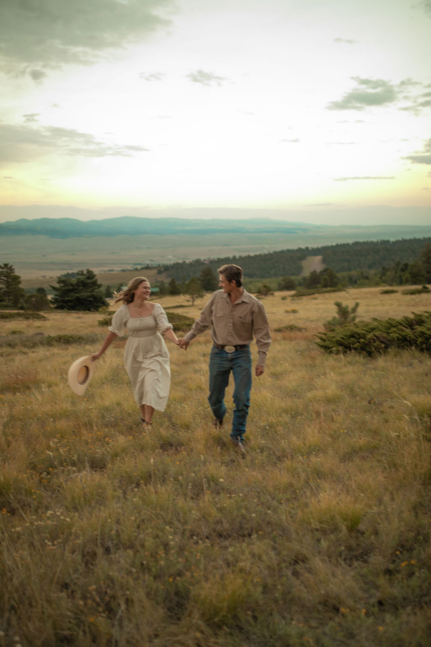 Couple holding hands walking through golden meadow with mountain valley sunset backdrop in rustic western attire.