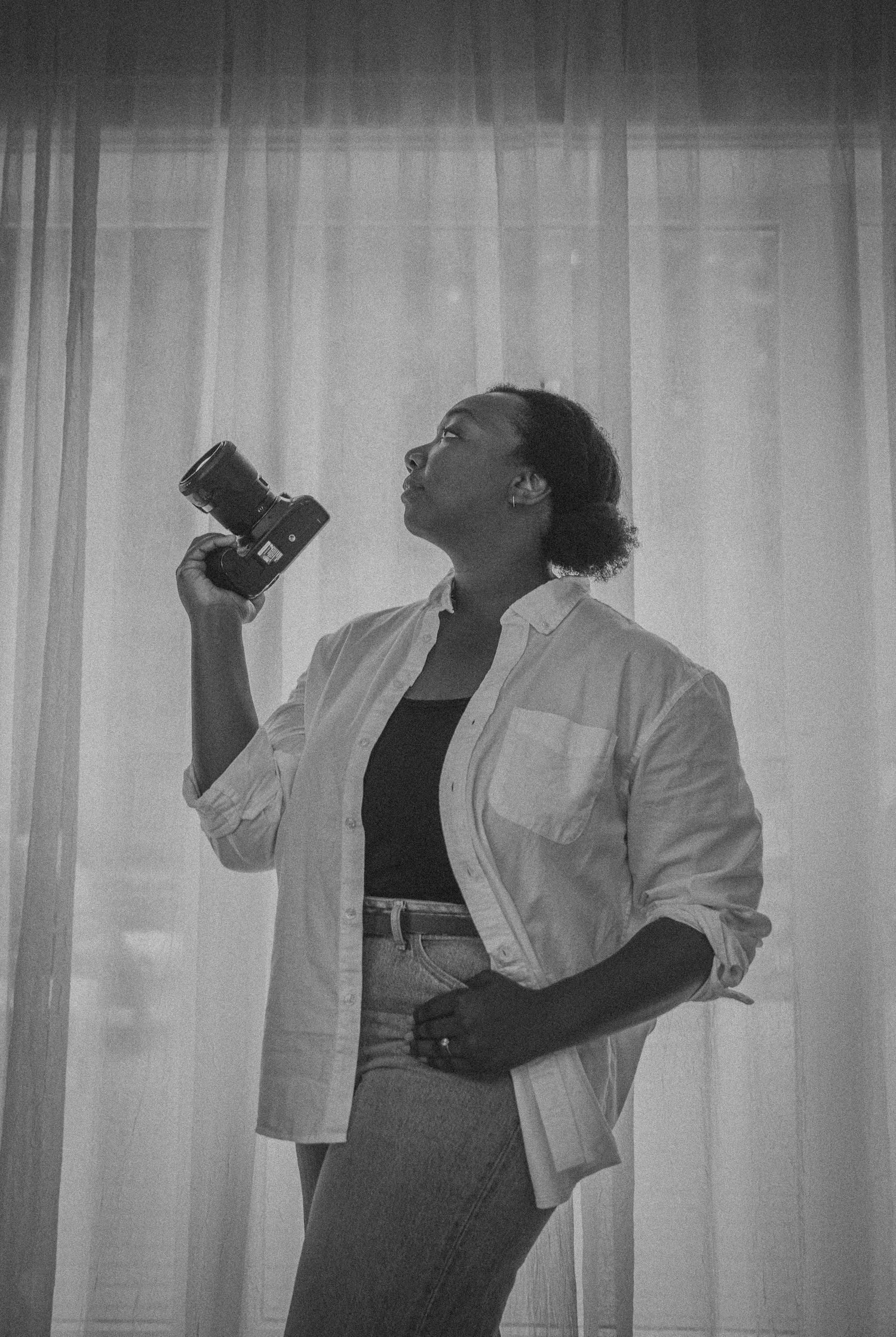 Black and white photo of a woman holding a camera up, wearing a white shirt, standing in front of sheer curtains.