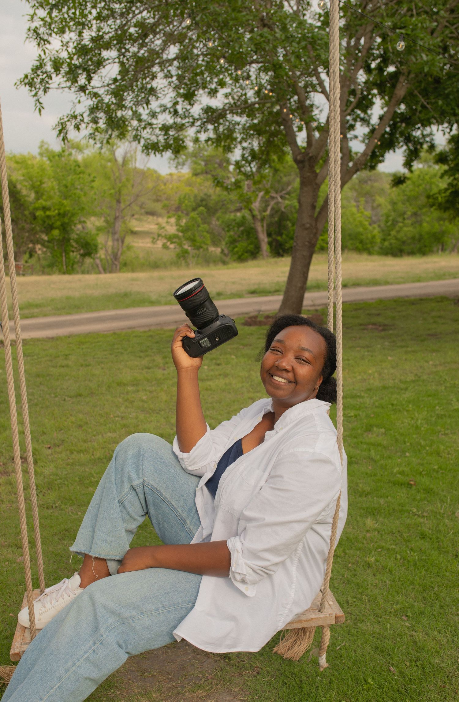 Smiling woman sitting on a rope swing holding a camera, wearing white shirt and jeans, with green trees in background.