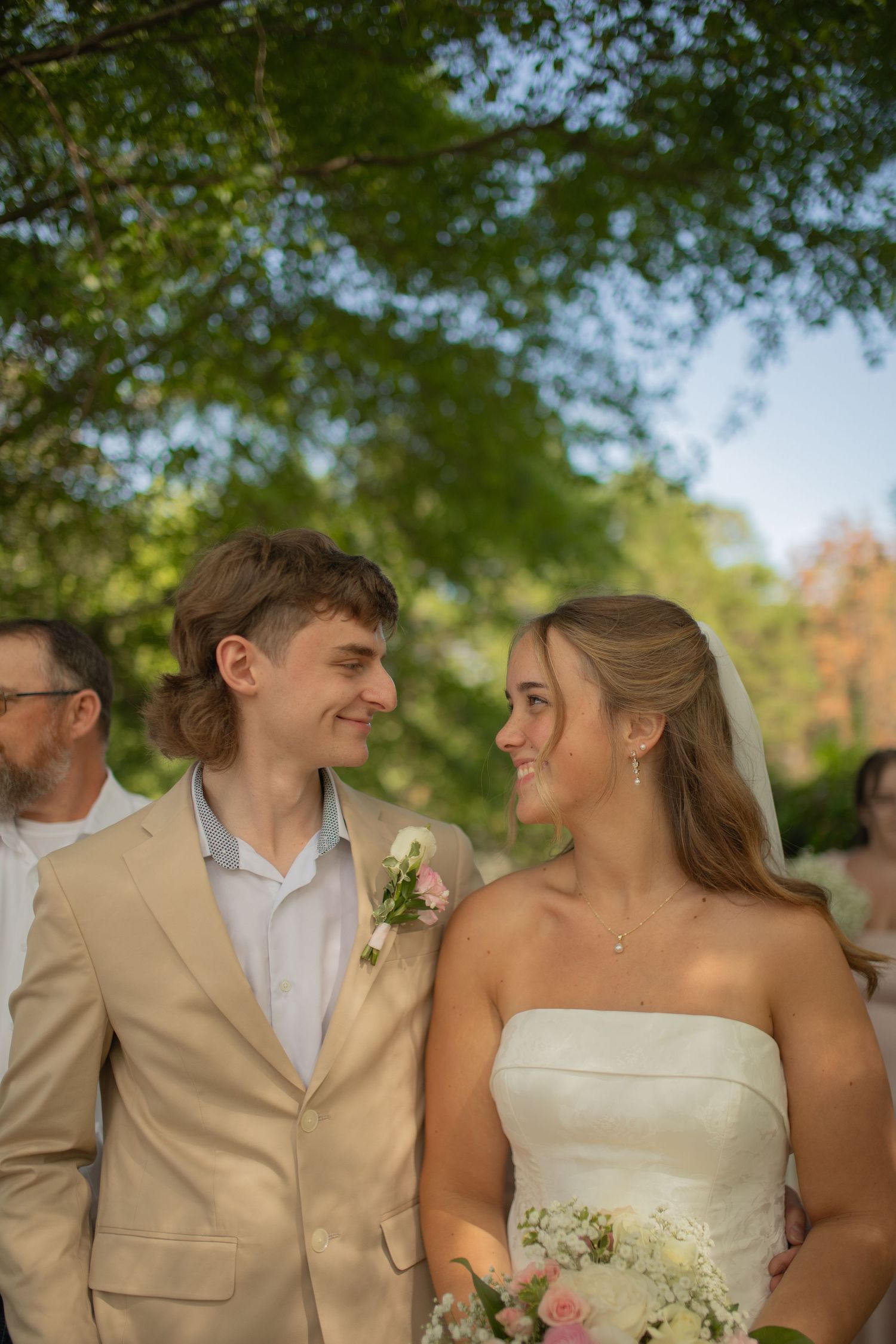 A young couple in wedding attire share a loving glance outdoors, groom in tan suit, bride in white gown holding pink flowers.