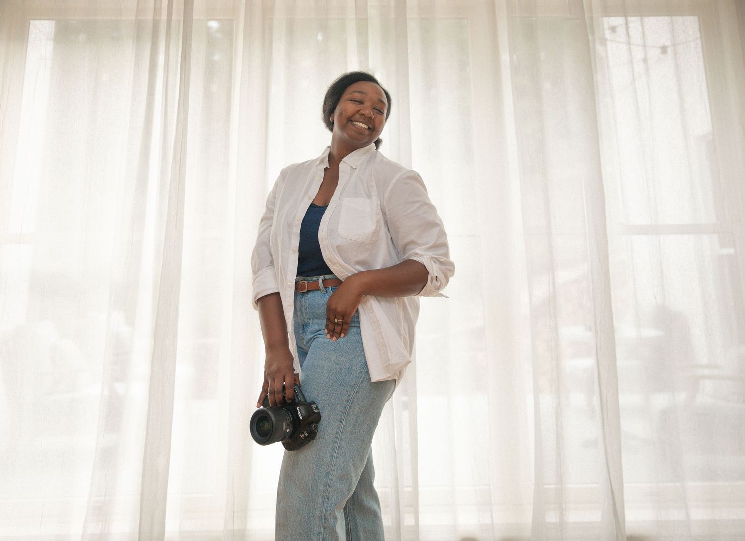 Smiling Black woman photographer holding a camera, wearing a white blazer and jeans, standing by sheer curtains.