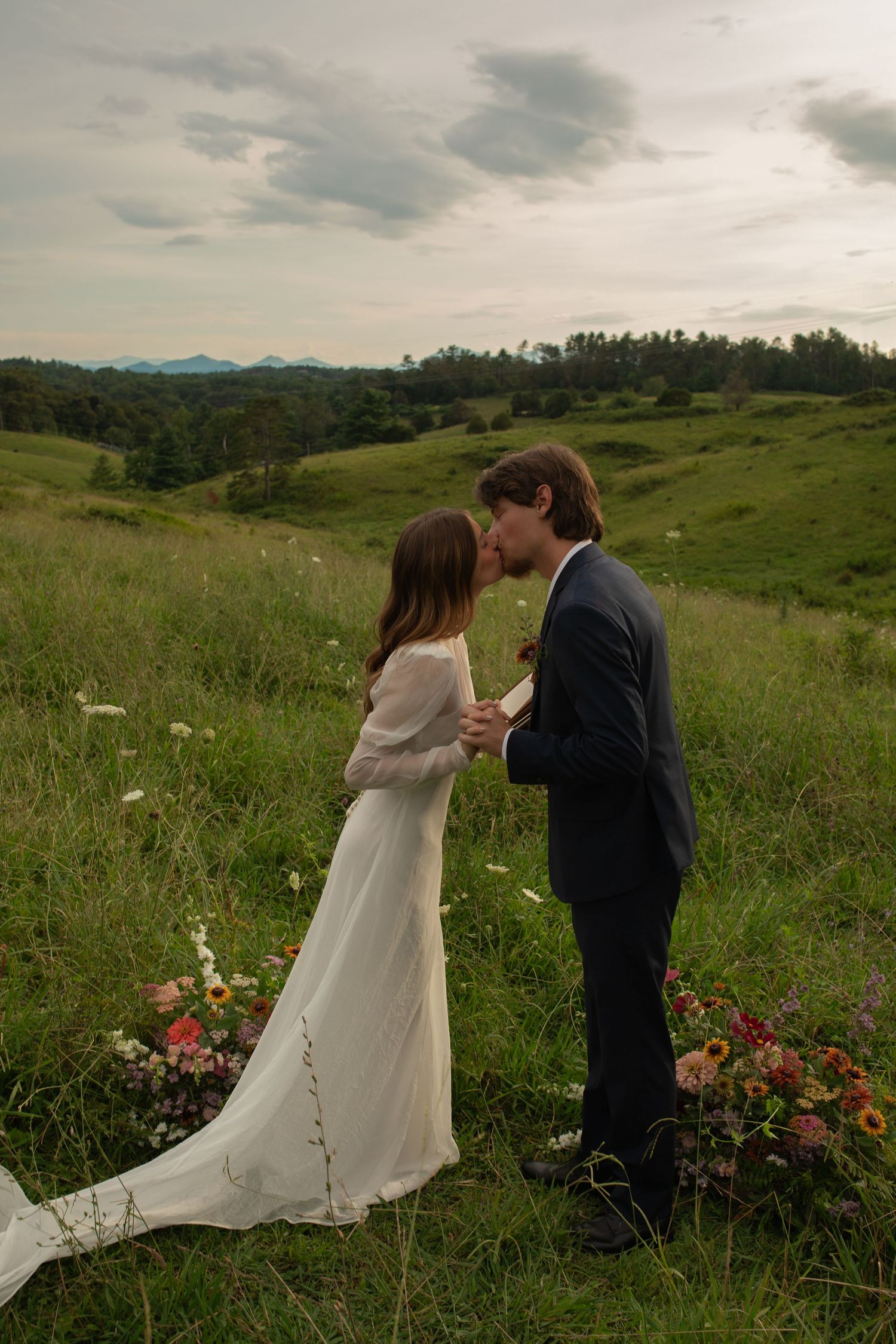 Bride and groom kissing in a lush green meadow, surrounded by wildflowers, with rolling hills and cloudy sky backdrop.