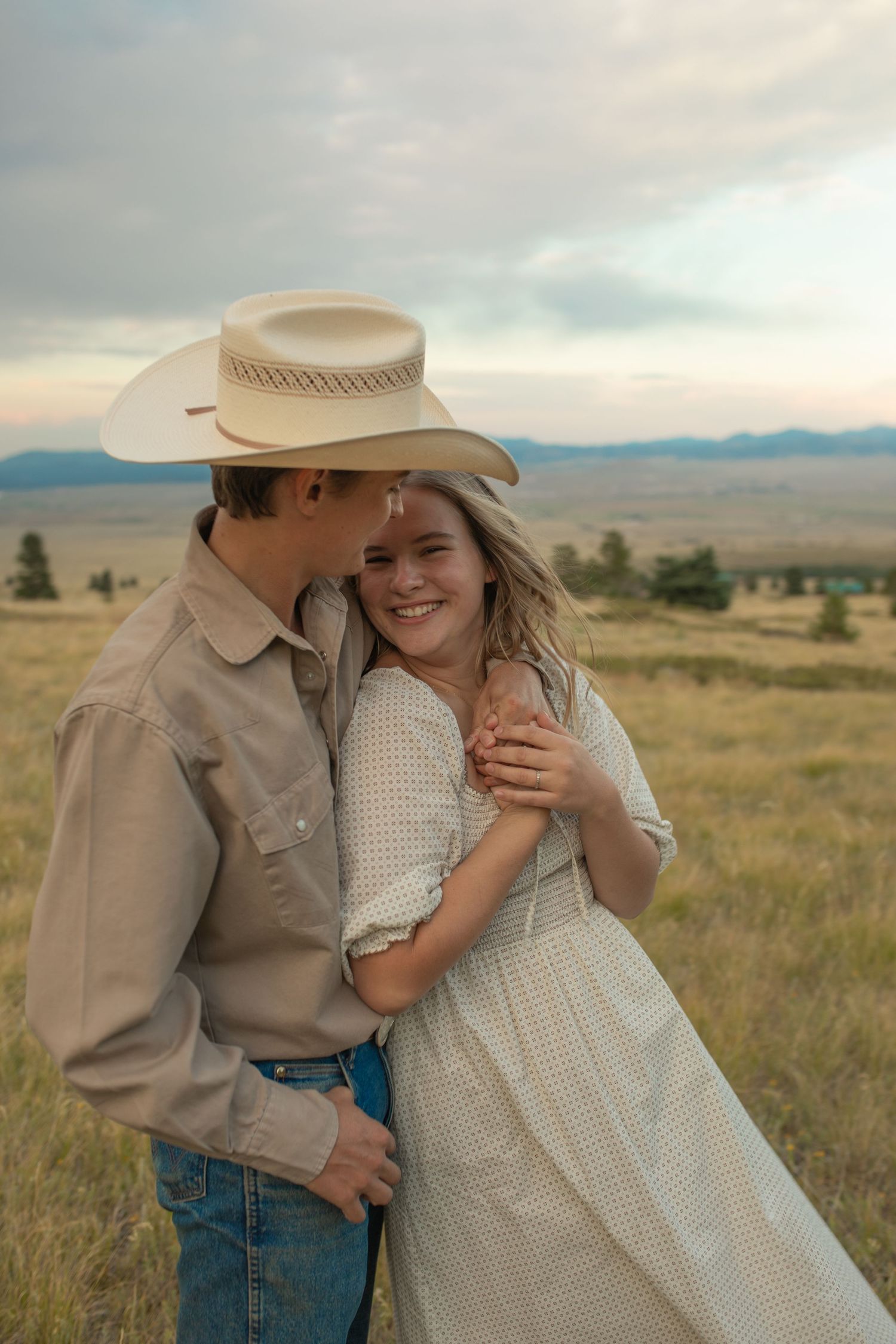 A young couple embraces in a golden field, the man in a cowboy hat kissing her cheek as she smiles.