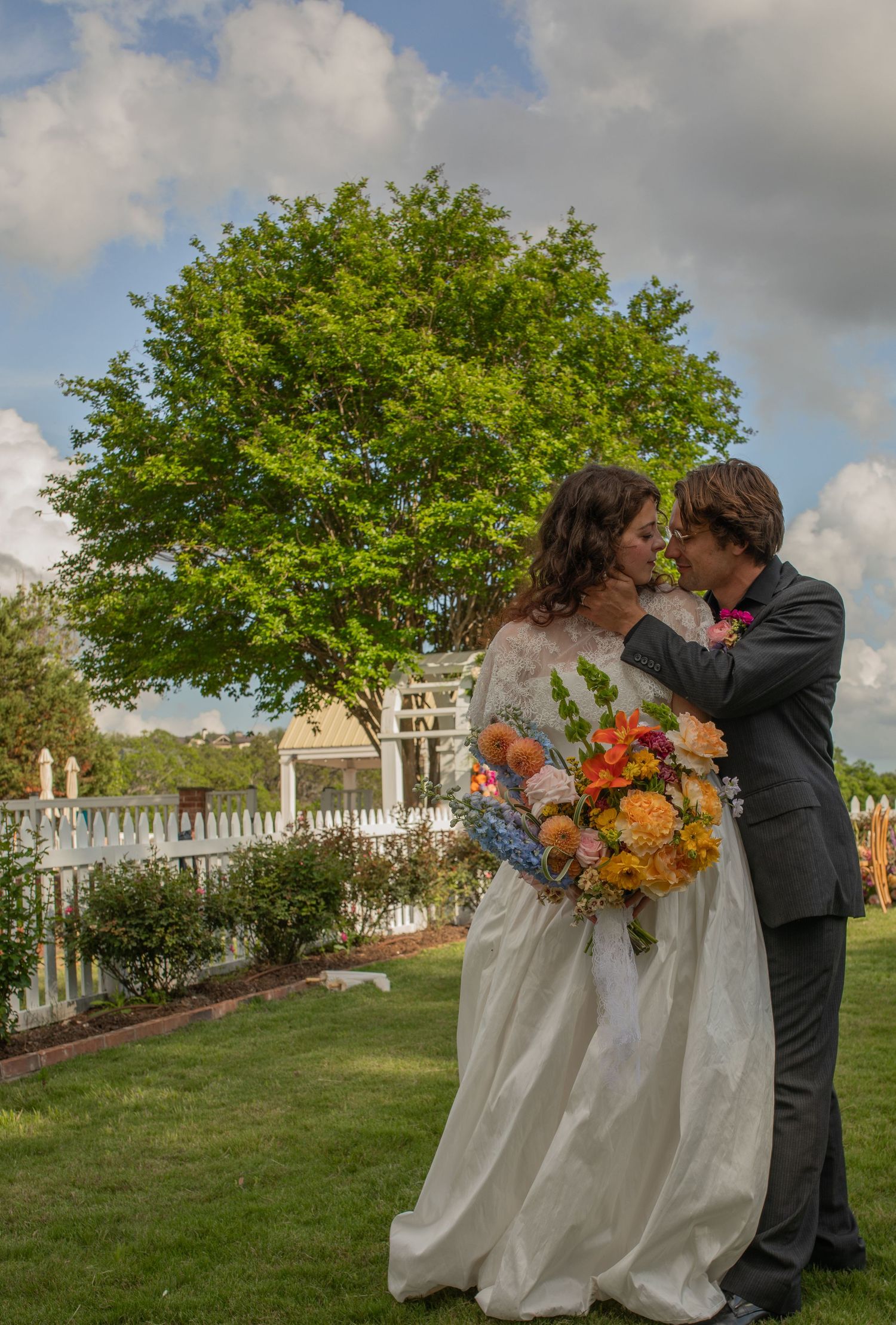 Bride and groom share a romantic kiss outdoors, bride holding colorful bouquet, lush green tree and white fence in background.