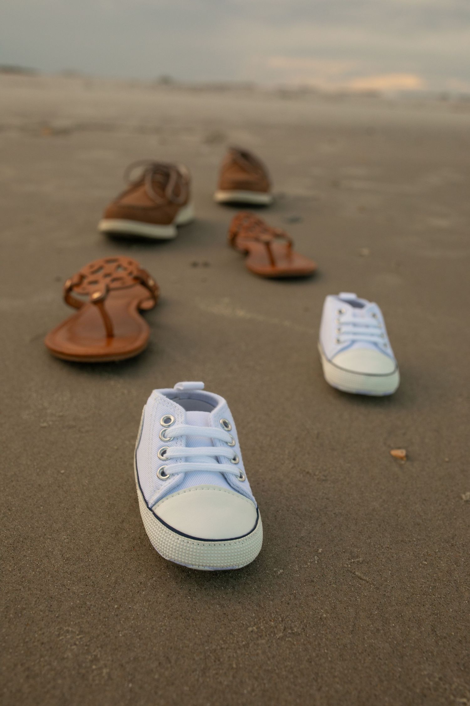 Various casual shoes including white sneakers and brown sandals arranged in a line on a sandy beach at sunset.