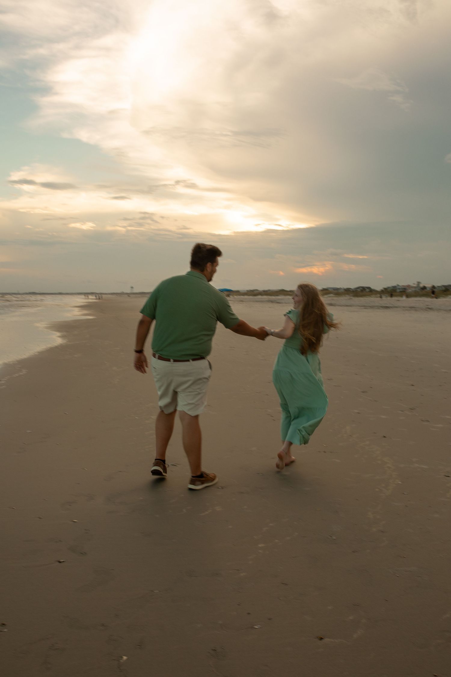 A couple holds hands while walking along a serene beach at sunset with cloudy skies overhead.