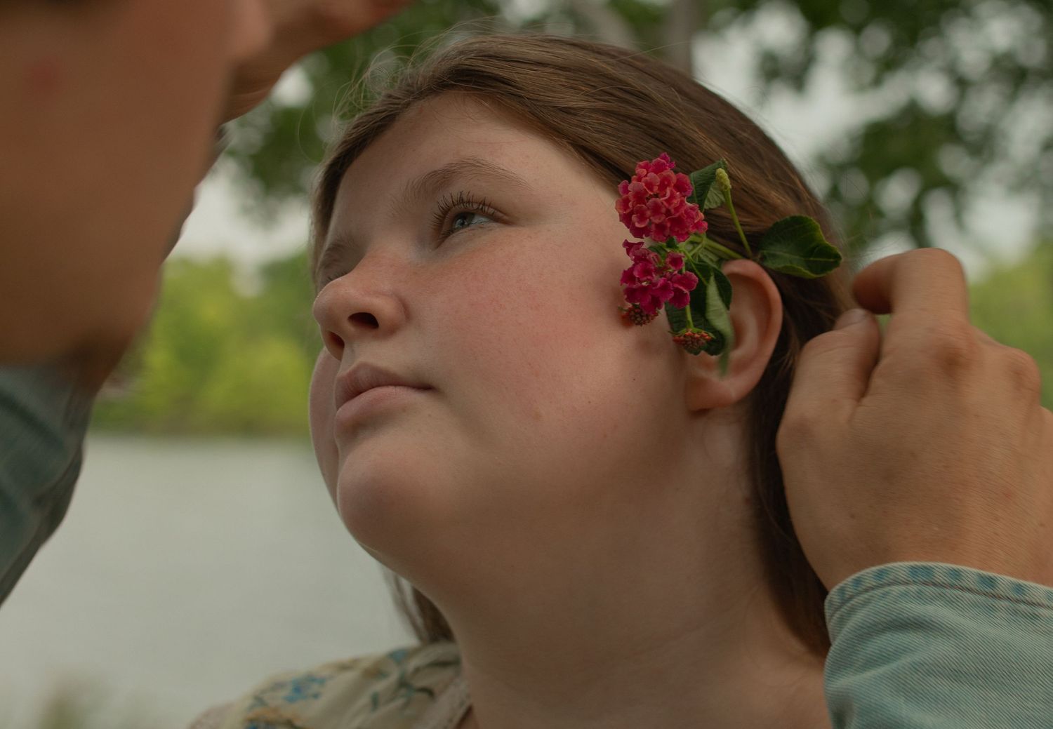 A child with red flowers tucked behind her ear by another person near a serene lakeside backdrop.