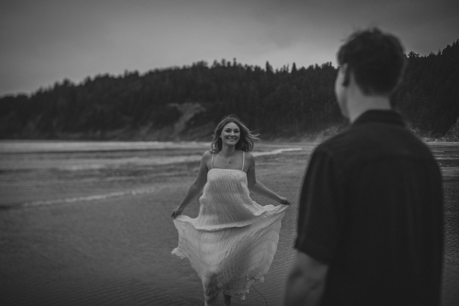 Black and white beach photo of a smiling woman in a flowing dress walking toward a man, with forested hills behind.