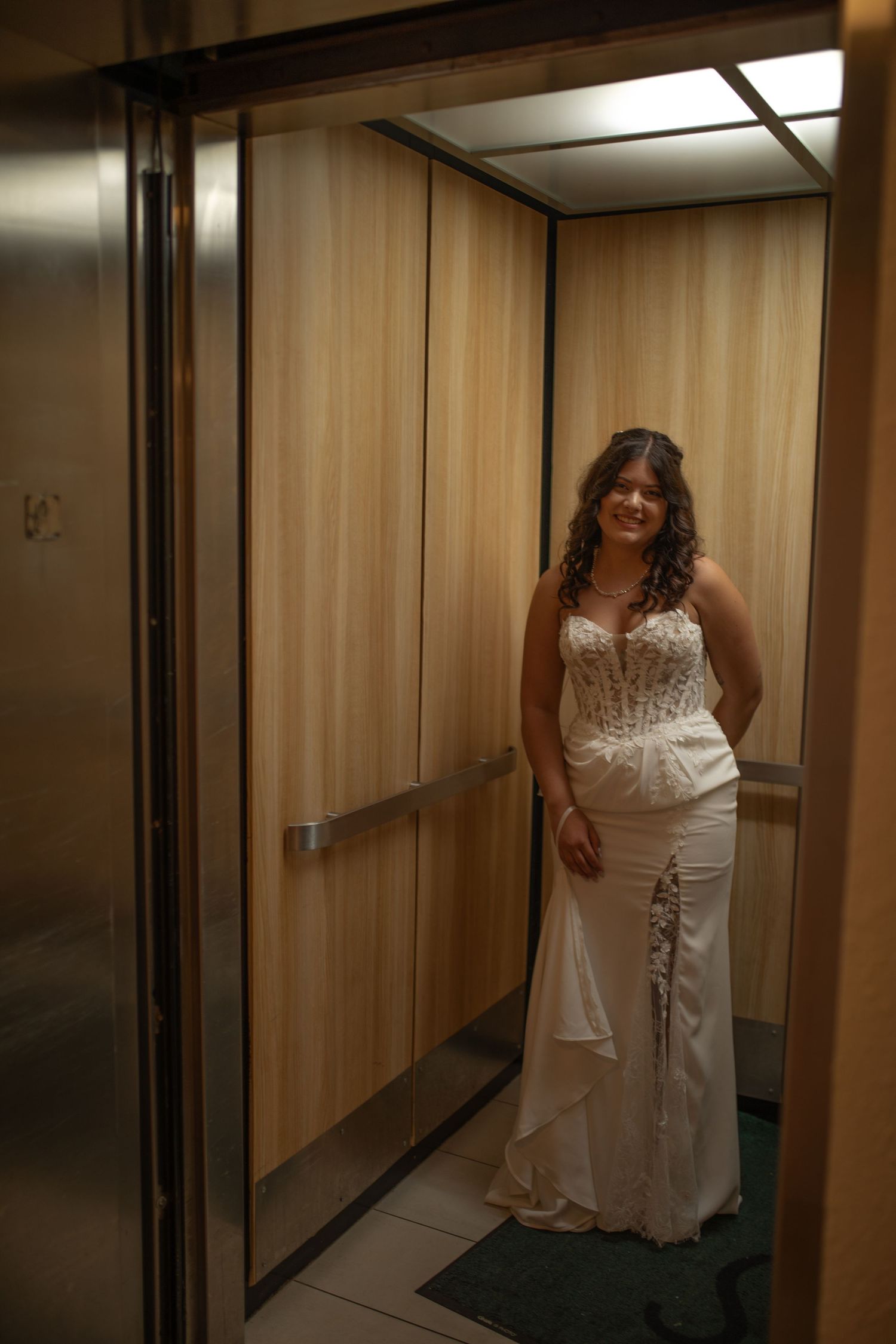 Bride in white wedding gown standing elegantly inside a wood-paneled elevator, smiling for the camera.