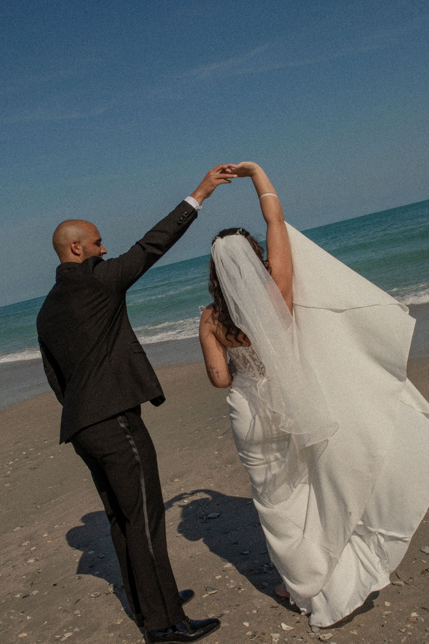 A groom in a black suit twirls his bride in a white gown and veil on a sunny beach wedding day.