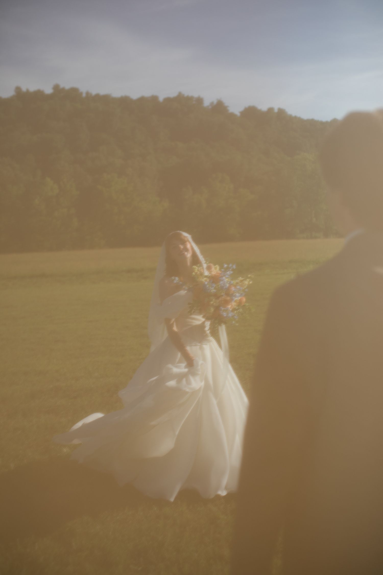 A bride in a flowing white gown and veil holds a wildflower bouquet in a sunlit meadow with trees in the background.