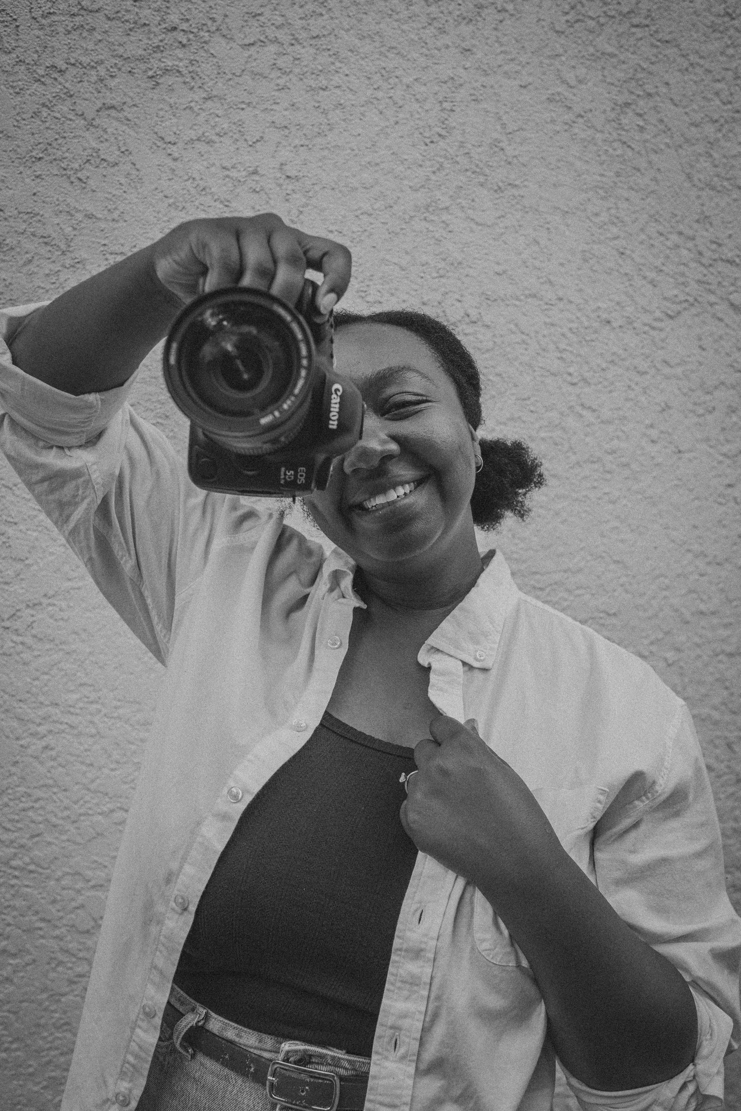Black and white photo of a smiling woman holding a Canon DSLR camera up to her face outdoors.