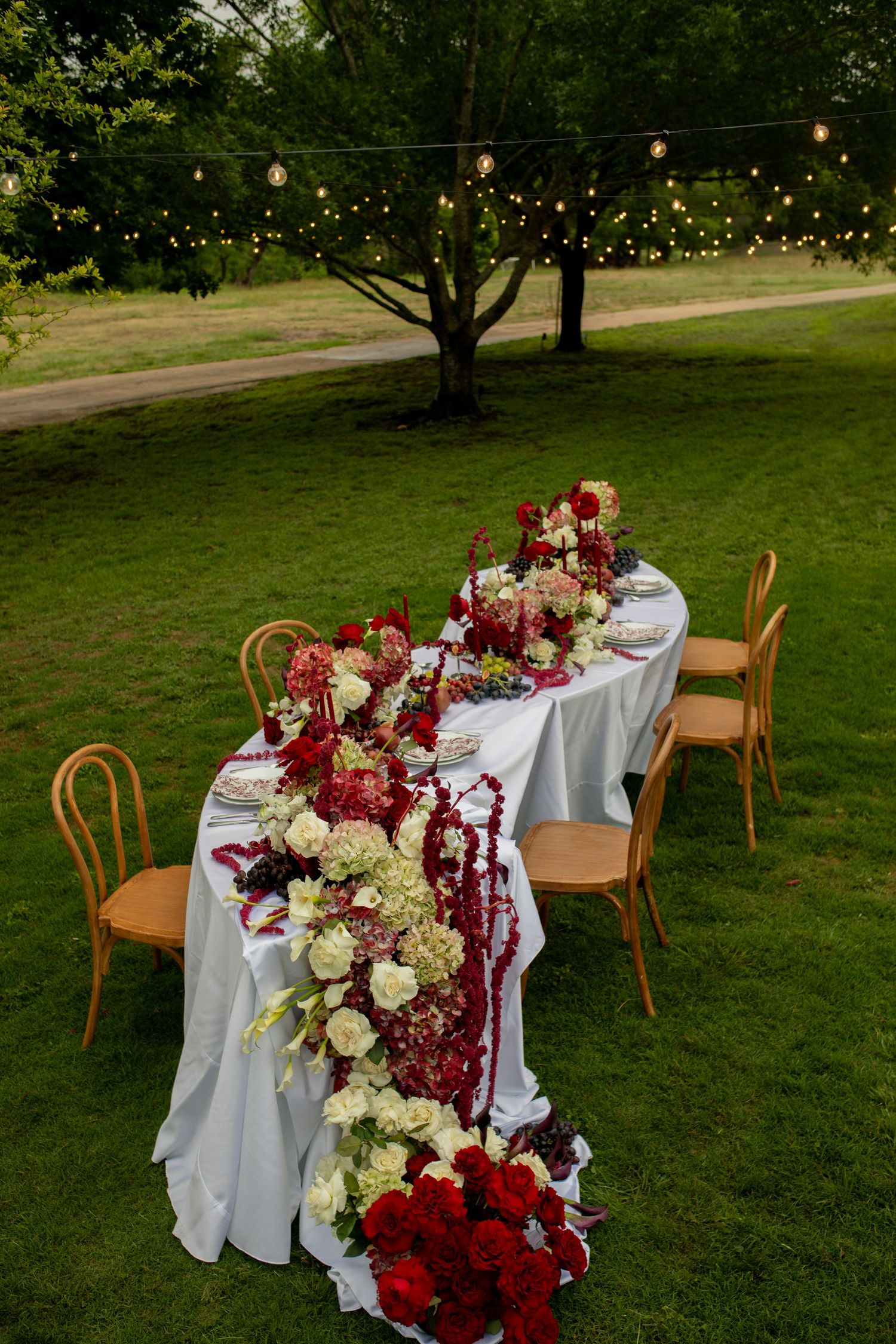 Elegant outdoor wedding reception table with red and white floral garland, string lights, and wooden chairs on lush green lawn.