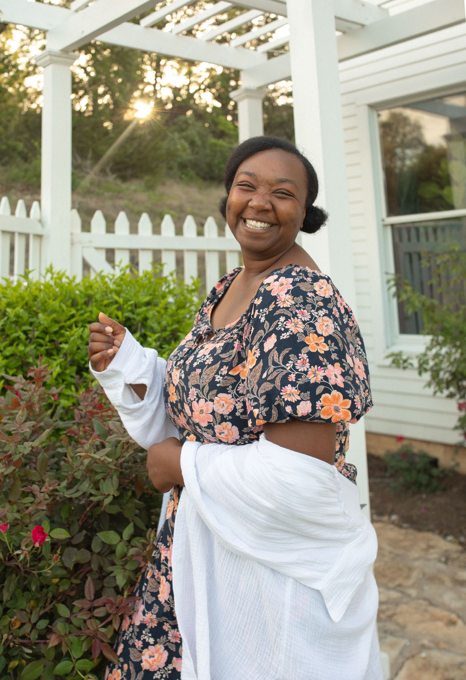 Smiling Black woman in floral crop top and white skirt posing outdoors near white pergola and garden.
