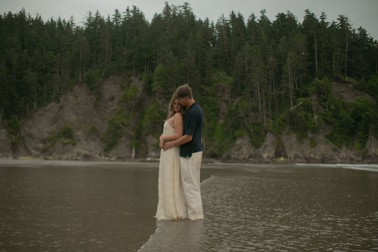Couple embracing on a misty beach with lush green forest cliffs in the background, romantic outdoor portrait.