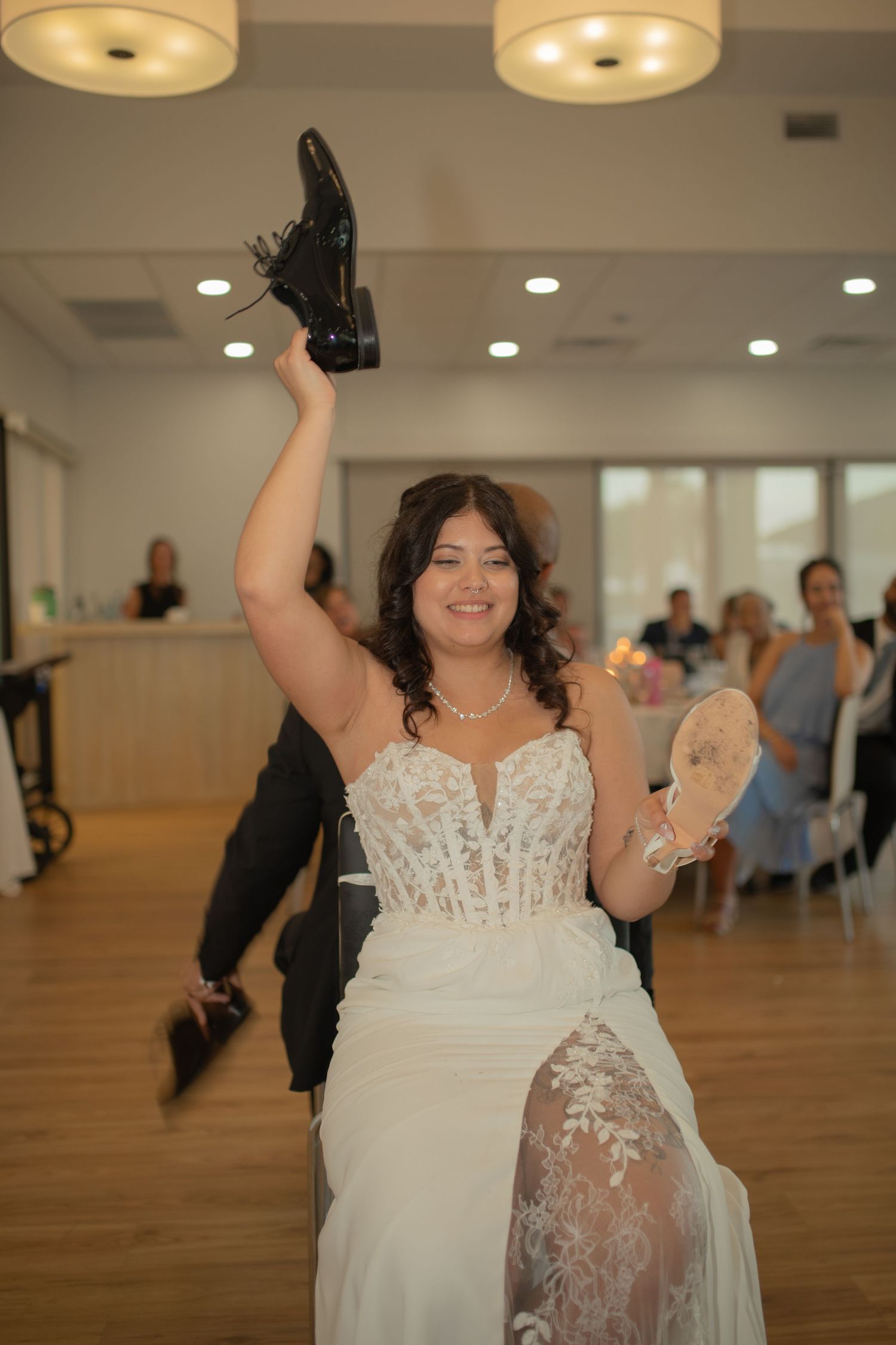 Smiling bride in white lace gown holds up a black shoe during a fun wedding reception game.