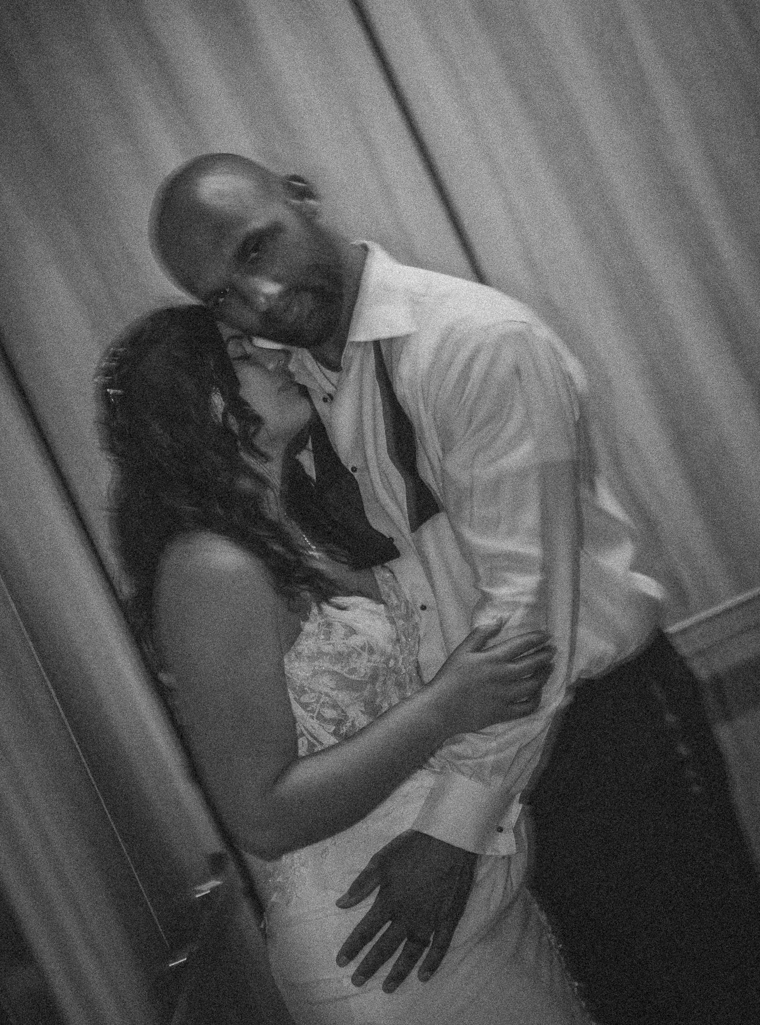 Black and white photo of a couple sharing a romantic kiss and embrace, possibly at a wedding reception.