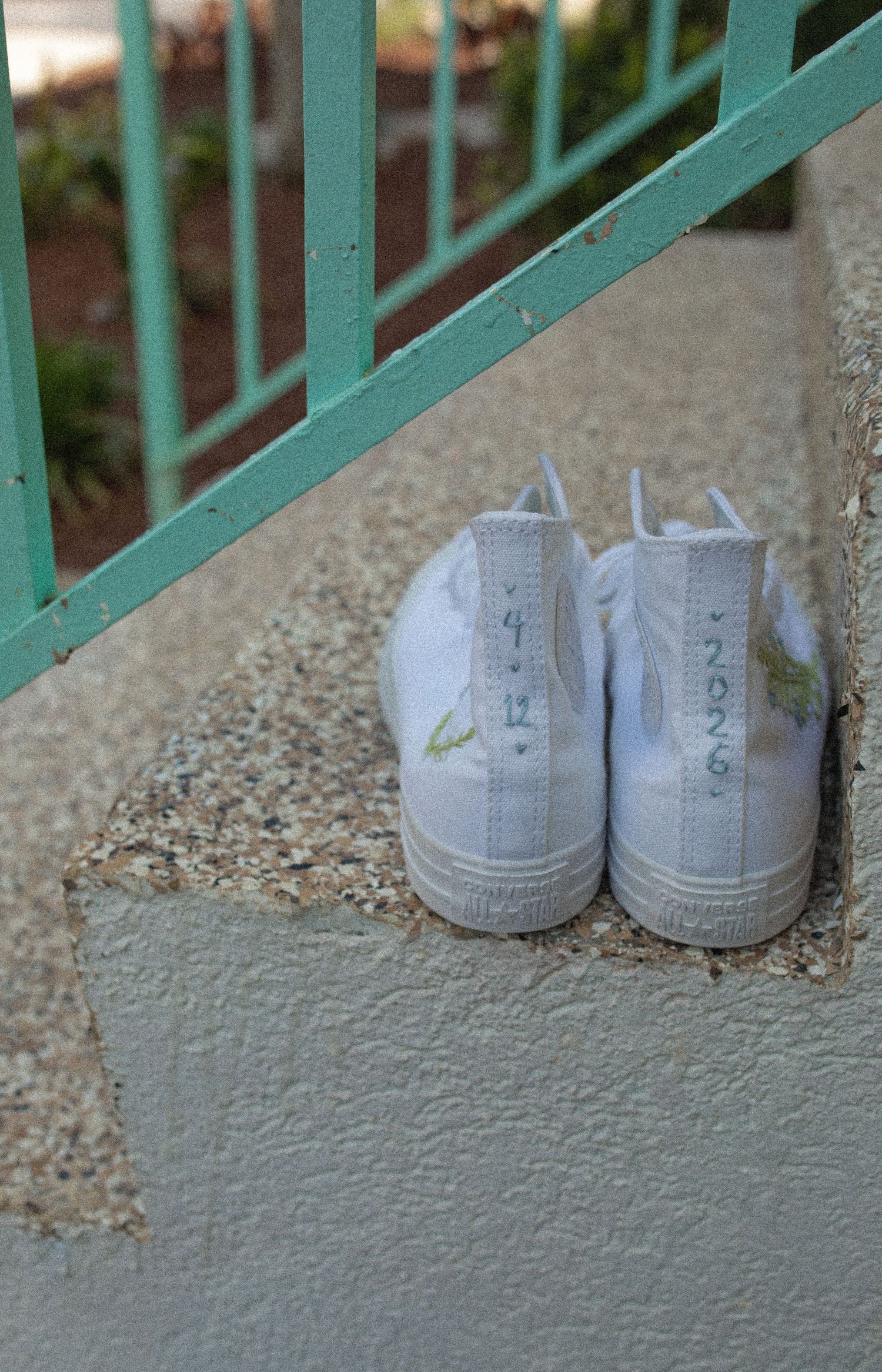 White high-top sneakers with green handwritten text sitting on concrete steps near a green metal railing.