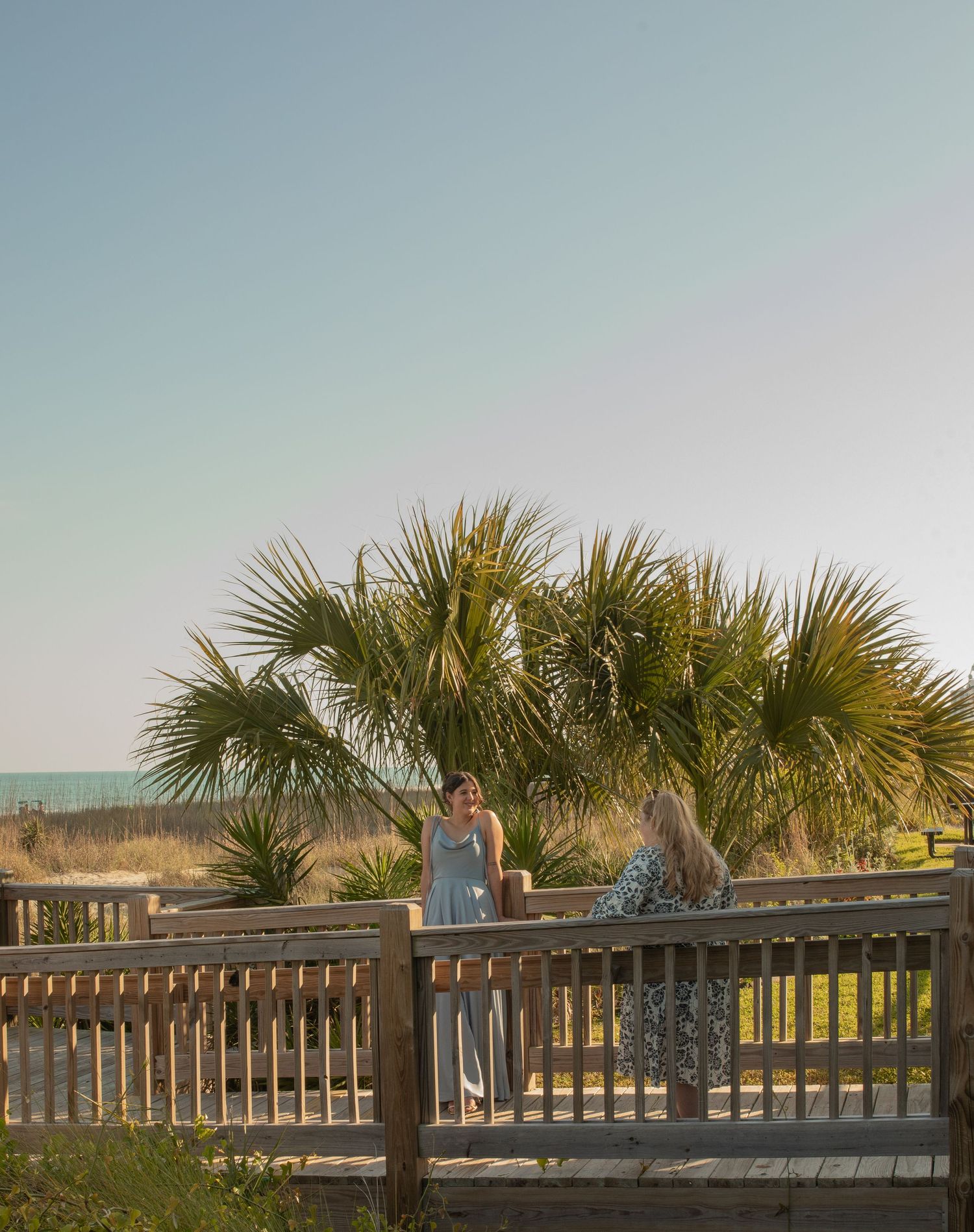 Two women chat on a wooden beachside deck surrounded by palm trees with ocean views at golden hour.
