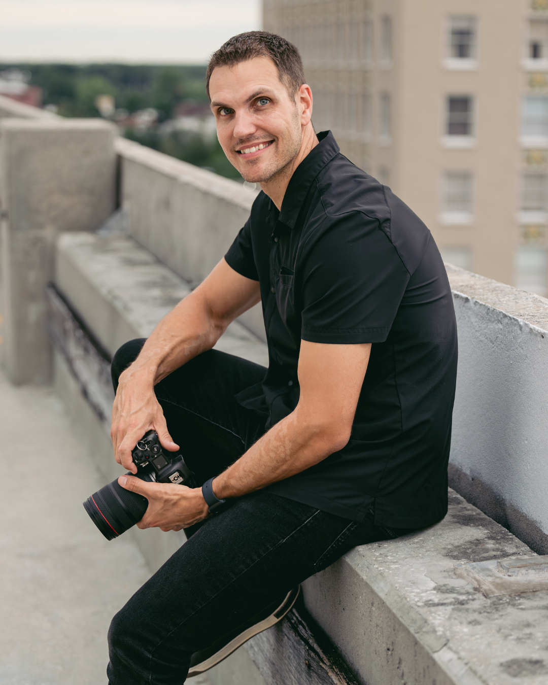 Nick Snell sitting on a ledge on a rooftop parking garage holding a canon camera in his hands smiling