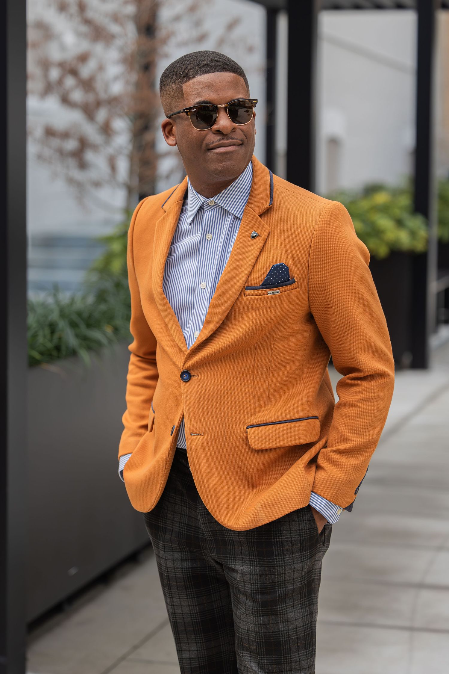Stylish man in bright orange blazer and plaid pants poses confidently outdoors.